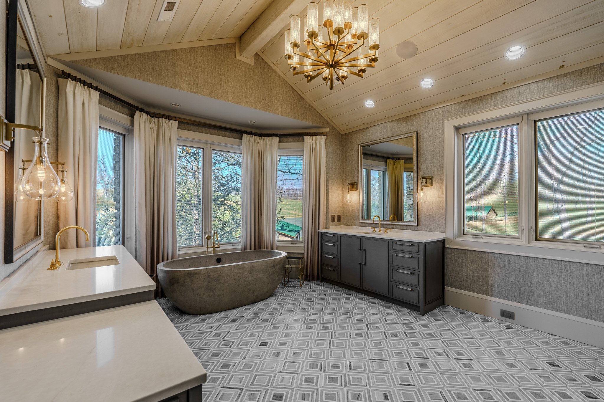 Modern bathroom with a freestanding bathtub near large windows, a dark gray vanity with a white marble countertop, and a large mirror above it. The ceiling is wood-paneled with a chandelier, and the floor features geometric patterned tile.