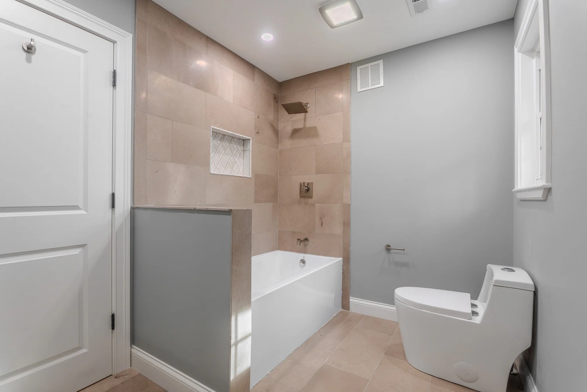 Modern bathroom with a bathtub, a toilet, gray wall, tile flooring, a small window, and a shower area with beige tiles.