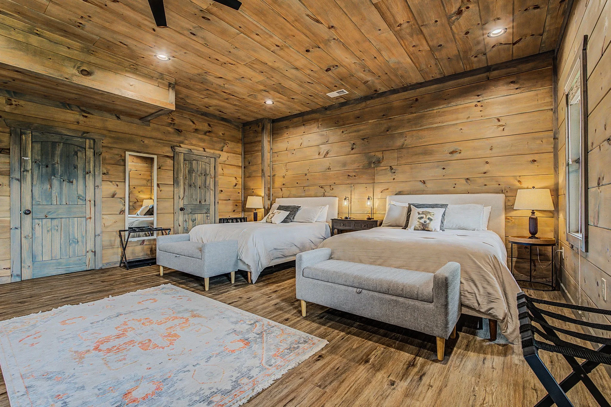 A bedroom with wooden walls and ceiling, two beds with white and grey bedding, a grey armchair, a white dresser, and a colorful area rug. There are lamps on the nightstands and a window with blinds.