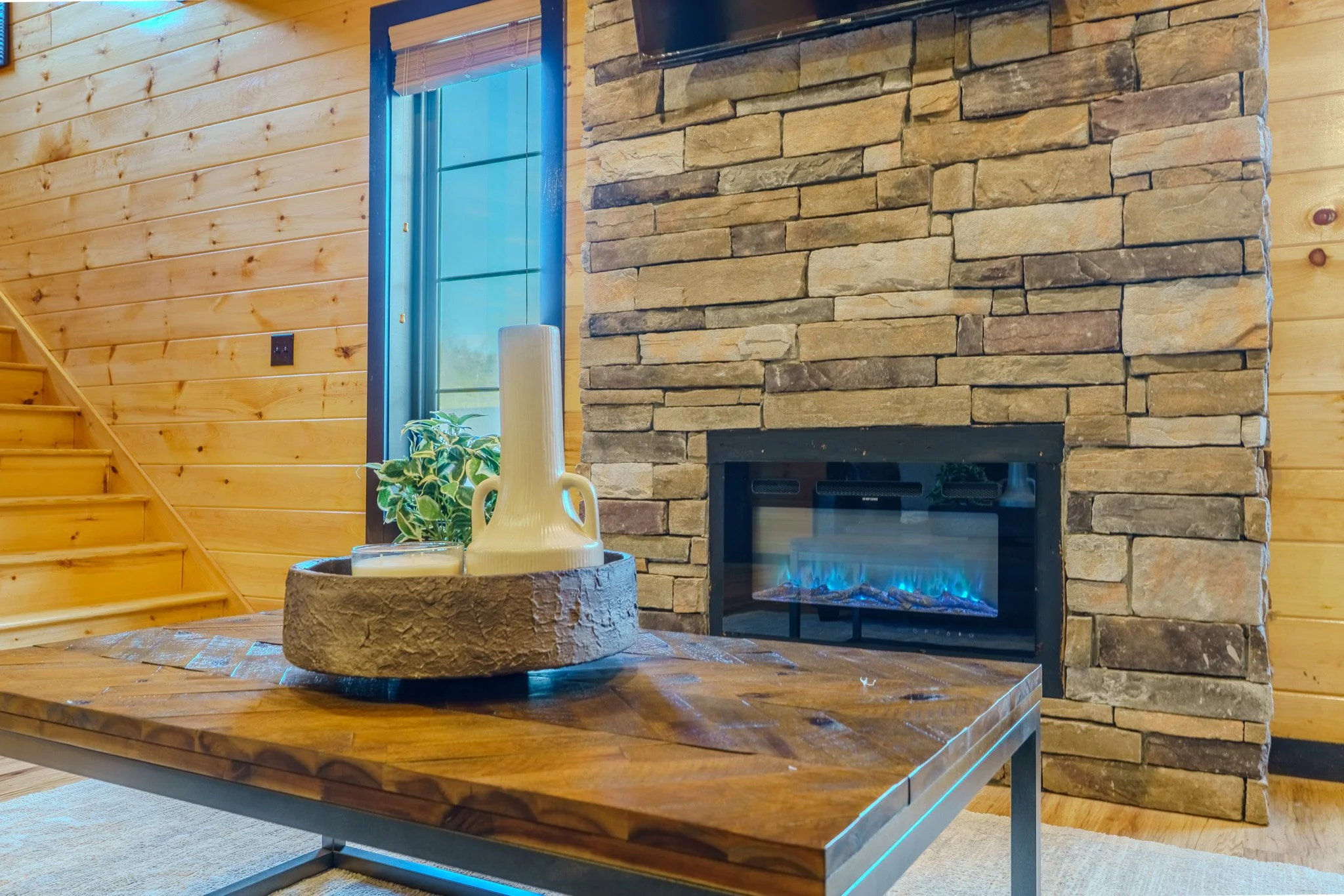 Living room with a wooden wall, stone fireplace with blue flame gas insert, wooden staircase, and a wooden coffee table with a decorative tray and plant.