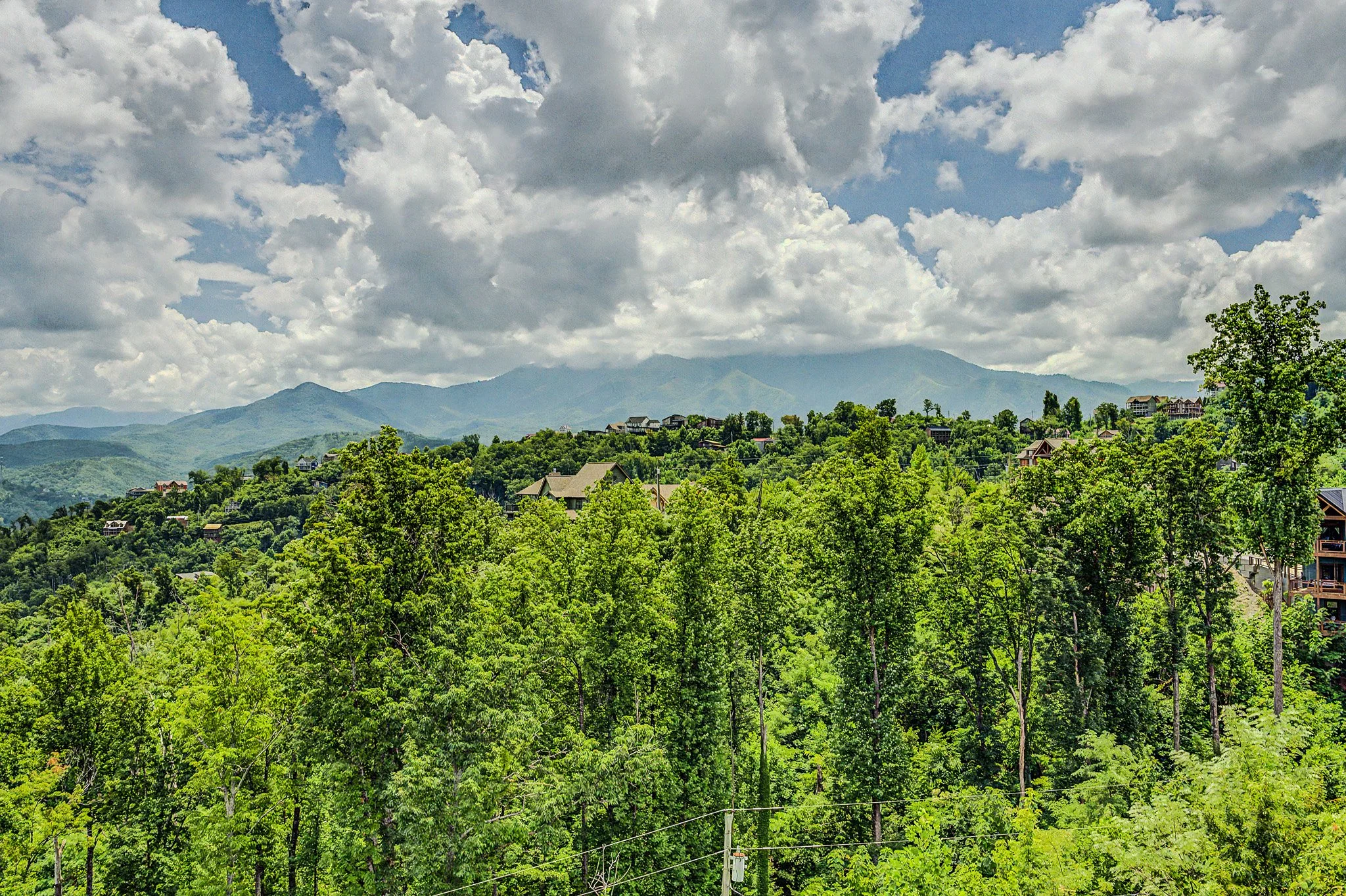 A scenic view of a mountain landscape with lush green trees in the foreground, houses on the hillside, and blue mountains in the background under a partly cloudy sky.