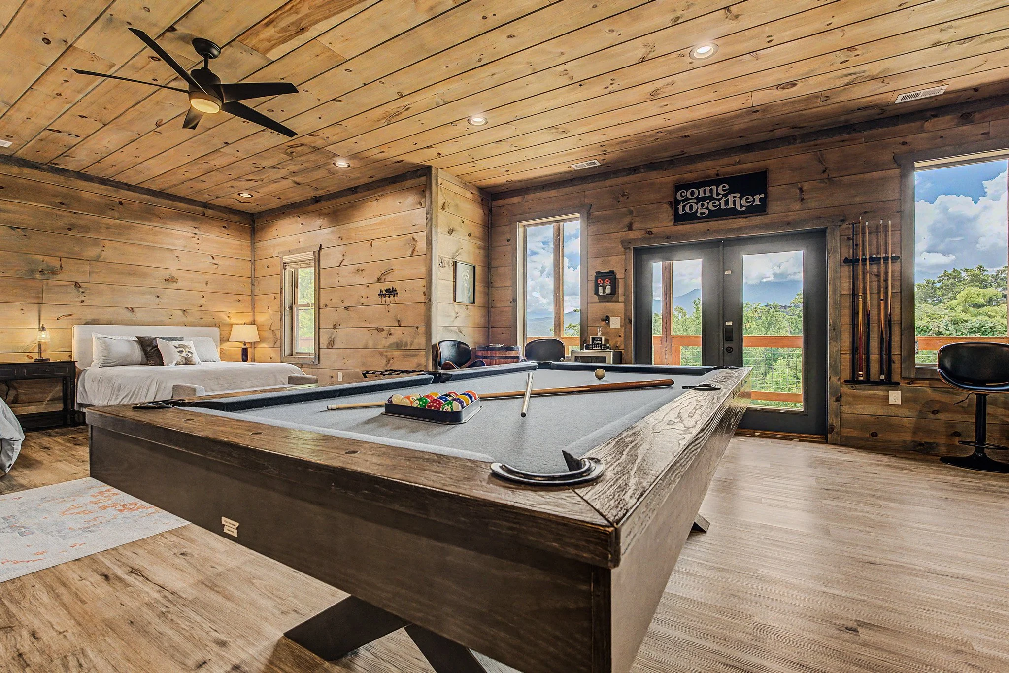 Interior of a rustic wooden cabin living space with a pool table, a bed, a nightstand with a lamp, and large windows showing a scenic mountain view.