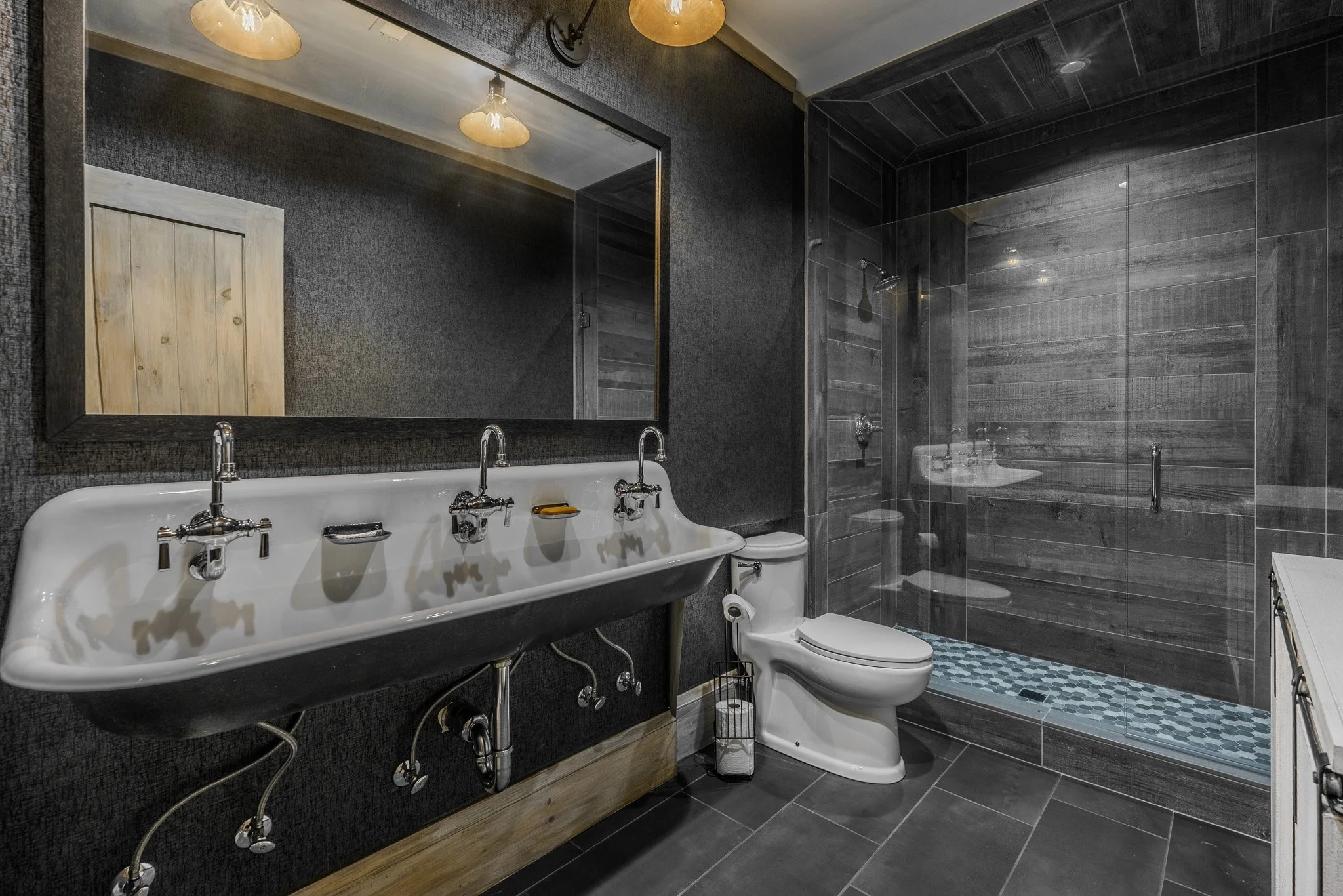 Modern bathroom with a sink, mirror, toilet, and walk-in shower with glass door. The walls and shower are tiled in dark gray with wood accents.