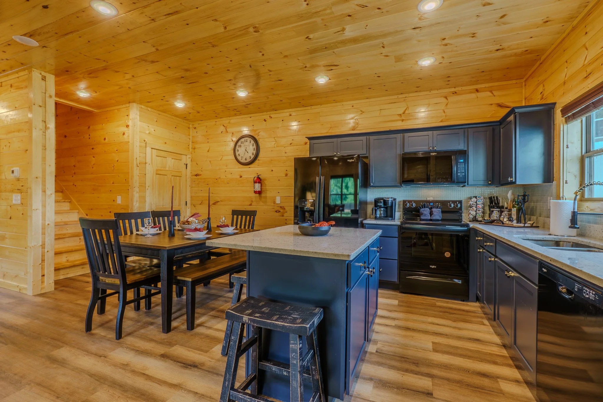 Kitchen with wooden walls and ceiling, black appliances, a central island with a bowl, a dining table set with wine glasses, a clock on the wall, and a window letting in natural light.
