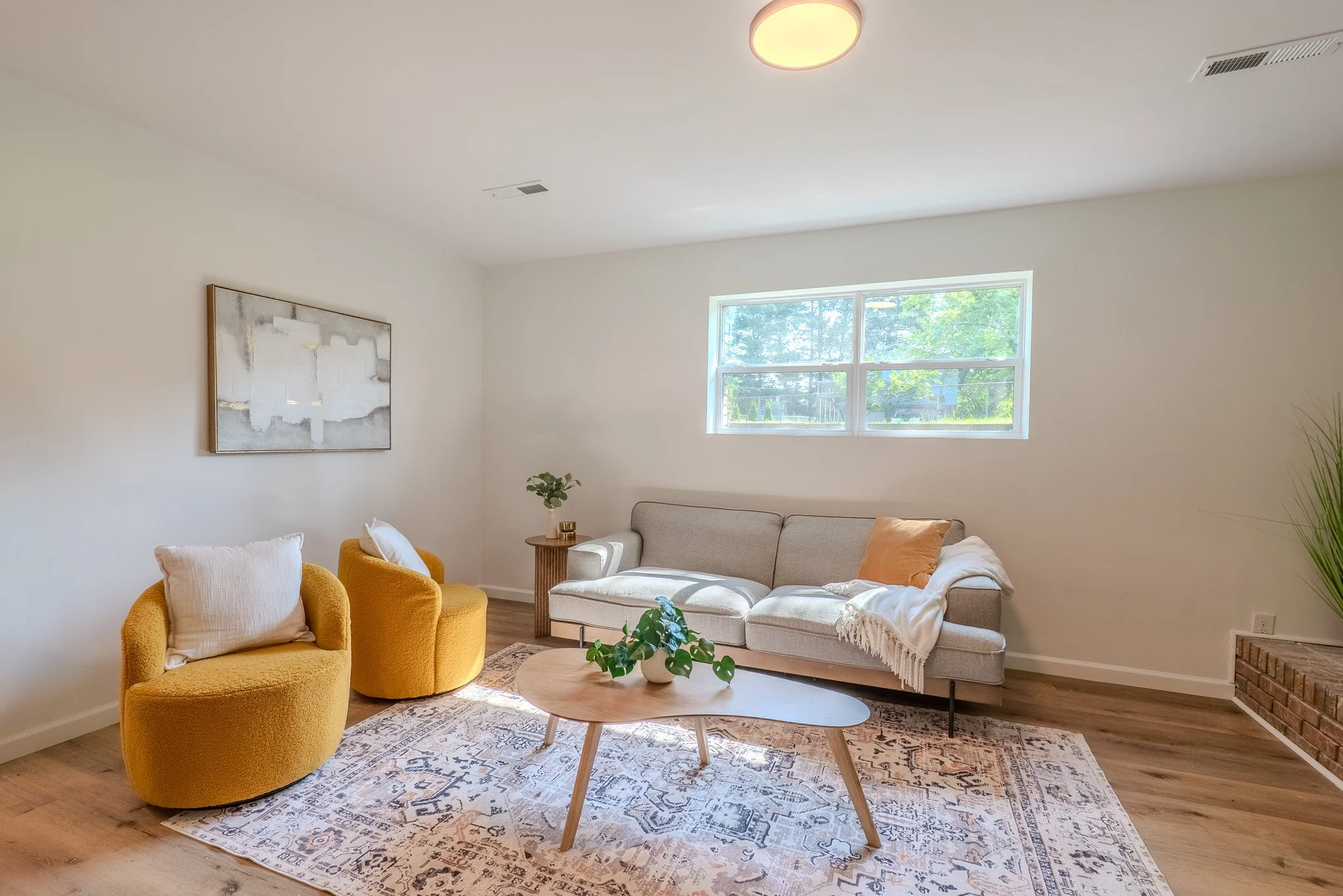 Minimalist living room with a beige sofa, two yellow armchairs, a wooden coffee table with a potted plant, a patterned rug, a window with trees outside, and wall art.