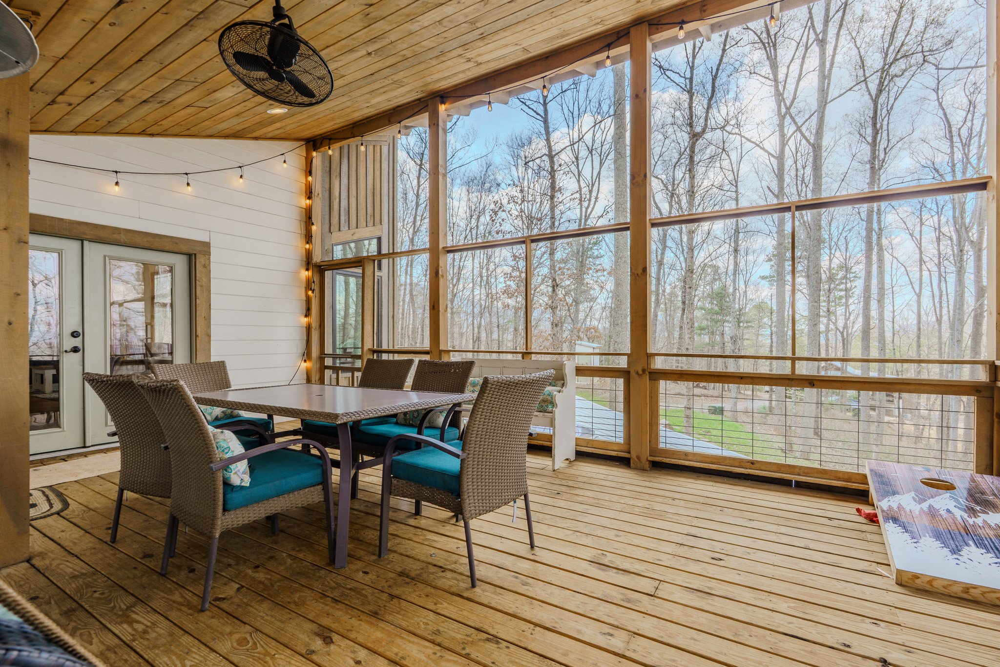An outdoor screened porch with wooden flooring, a dining table with six chairs, string lights, and a view of leafless trees outside.