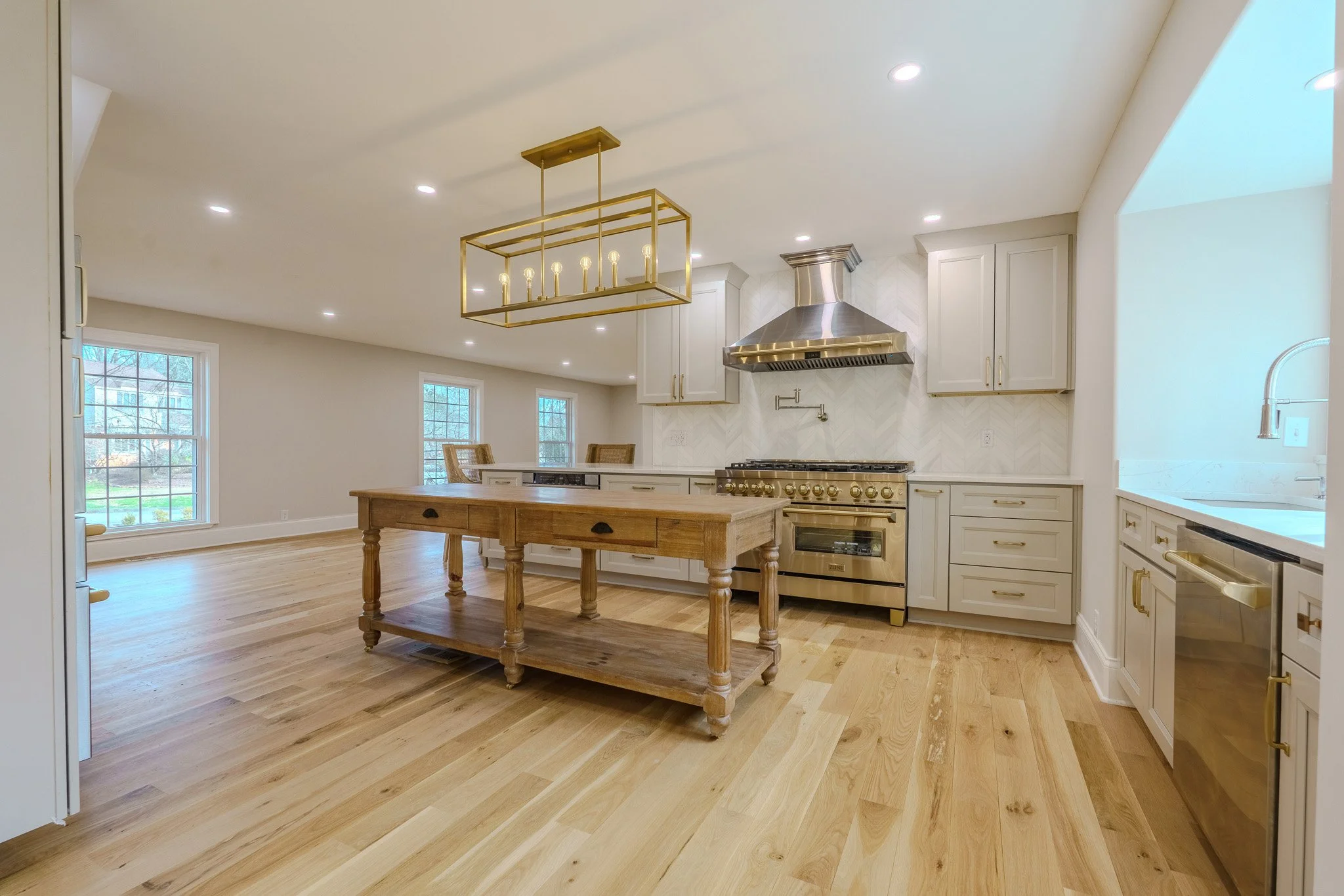 Open kitchen with white cabinetry, gold hardware, a stainless steel range with a gold oven, stainless steel vent hood, and a wooden kitchen island. Large windows letting in natural light, wood flooring, and a modern gold chandelier overhead.