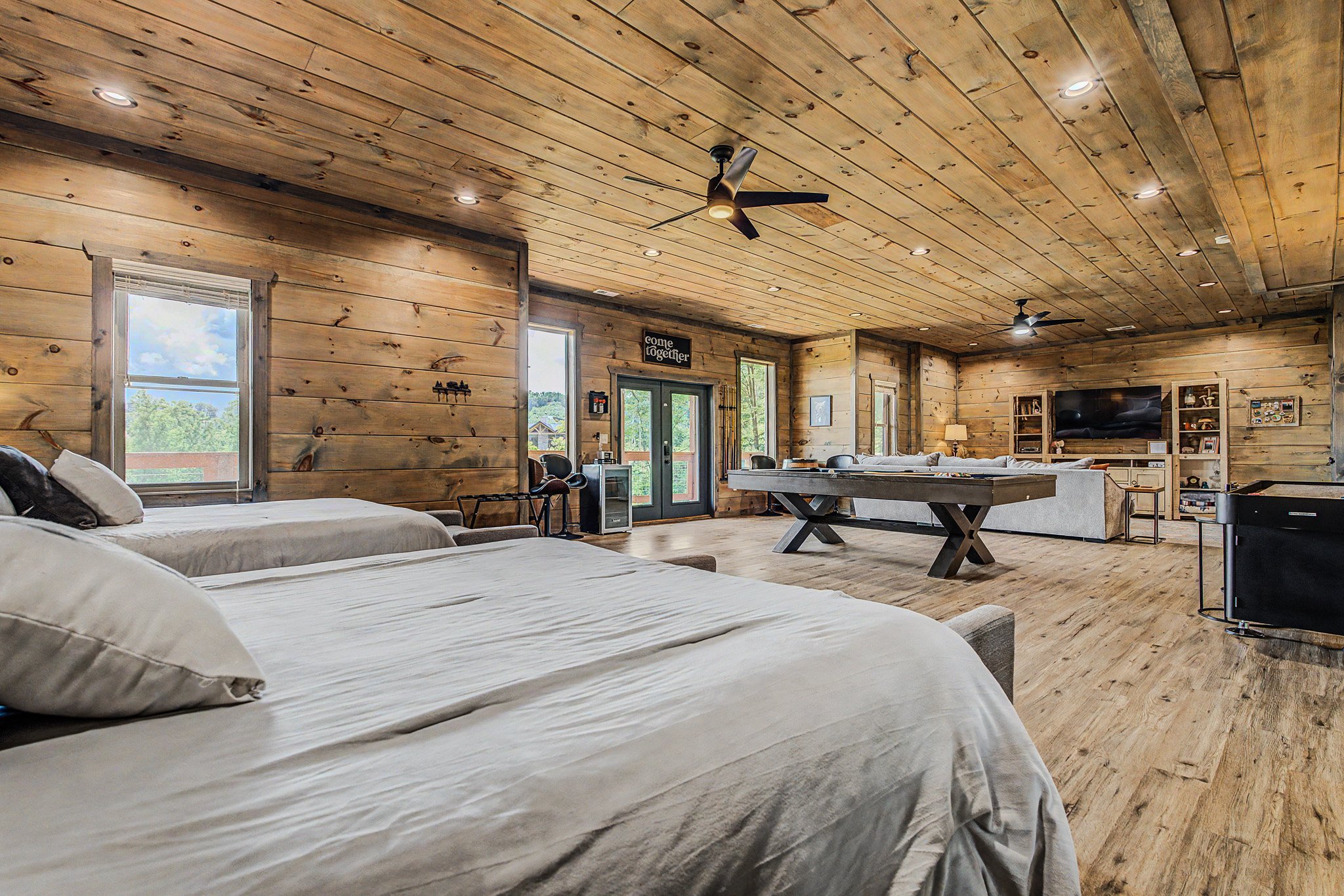 A spacious living area with wooden walls and ceiling, including two ceiling fans and multiple windows letting in natural light. There are beds with white linens in the foreground, a large flat-screen TV on the wall, and a dining table in the middle o