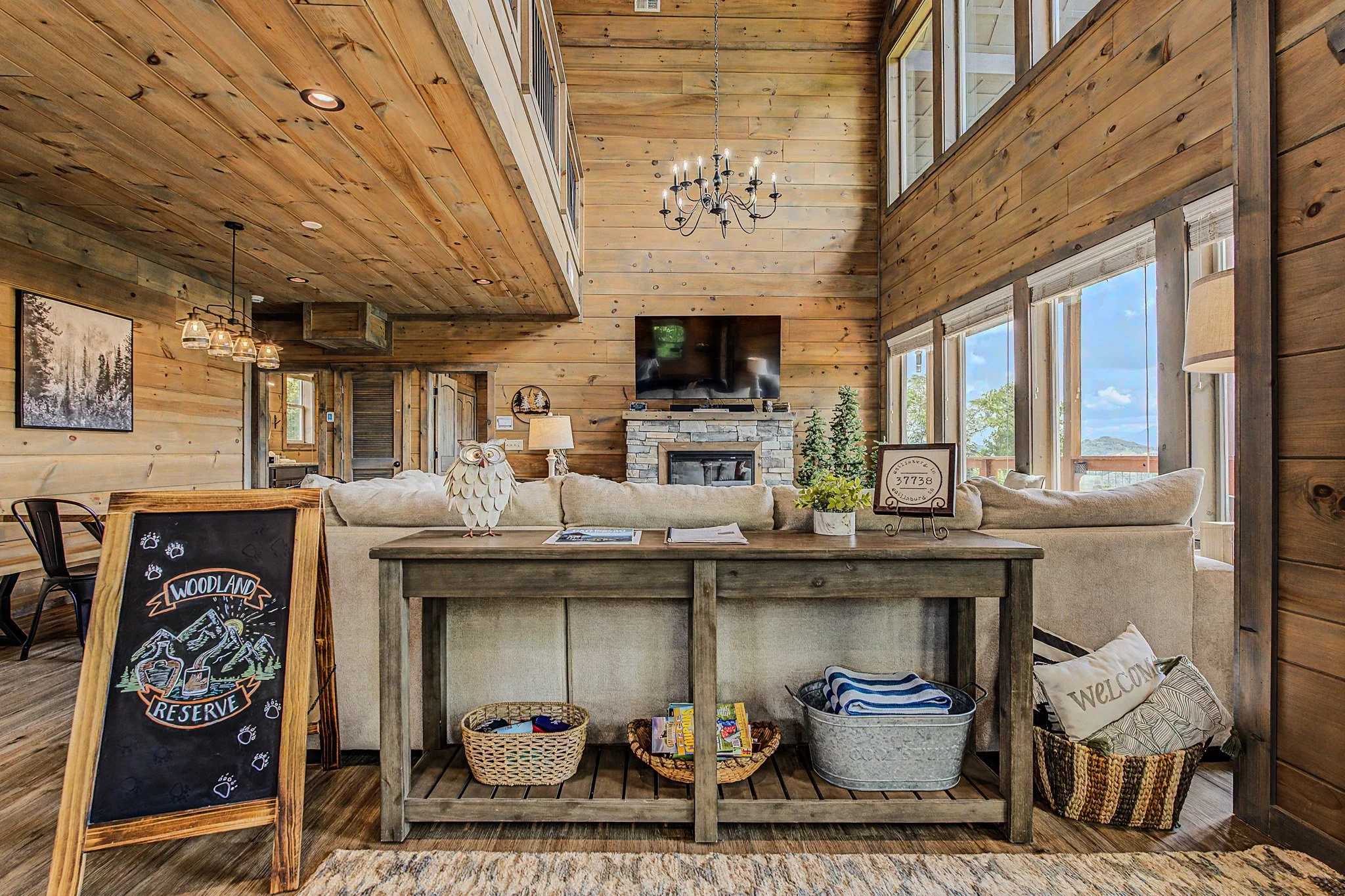 Interior of a rustic living room with wood-paneled walls and ceiling, large windows, a chandelier, a flat-screen TV above a stone fireplace, a beige sofa, various decorations, and a chalkboard with a mountain-themed drawing and text.
