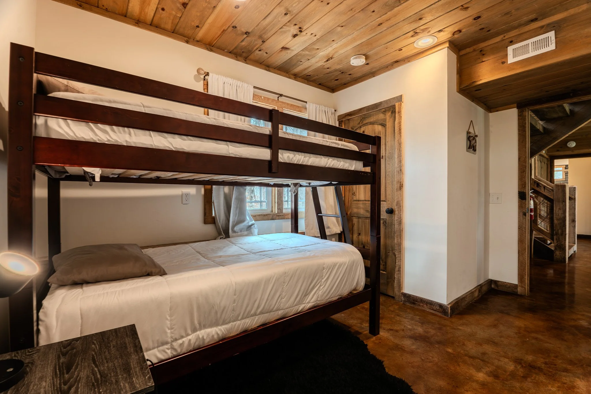 A rustic bedroom featuring a bunk bed with a single mattress on the top and a larger mattress on the bottom. The room has wooden ceiling panels, a window with white curtains, and a wooden door. The floor appears to be stained concrete or wood, and th