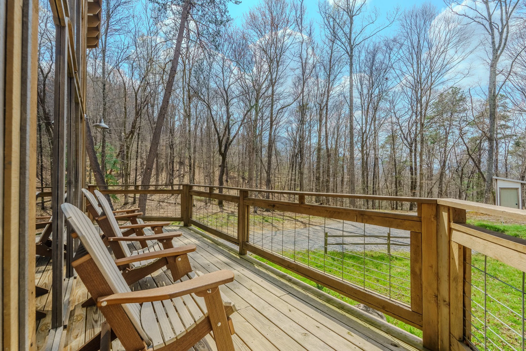 View of a wooden deck with four wooden chairs overlooking a wooded forest area with tall leafless trees during early spring or late fall, under a partly cloudy sky.