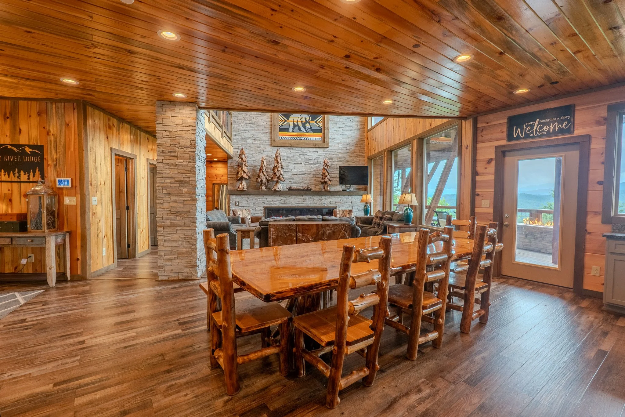 A rustic living and dining area with wooden walls and ceiling, a large central wooden dining table with log-style chairs, a stone fireplace with a TV above, and large windows showing an outdoor view.