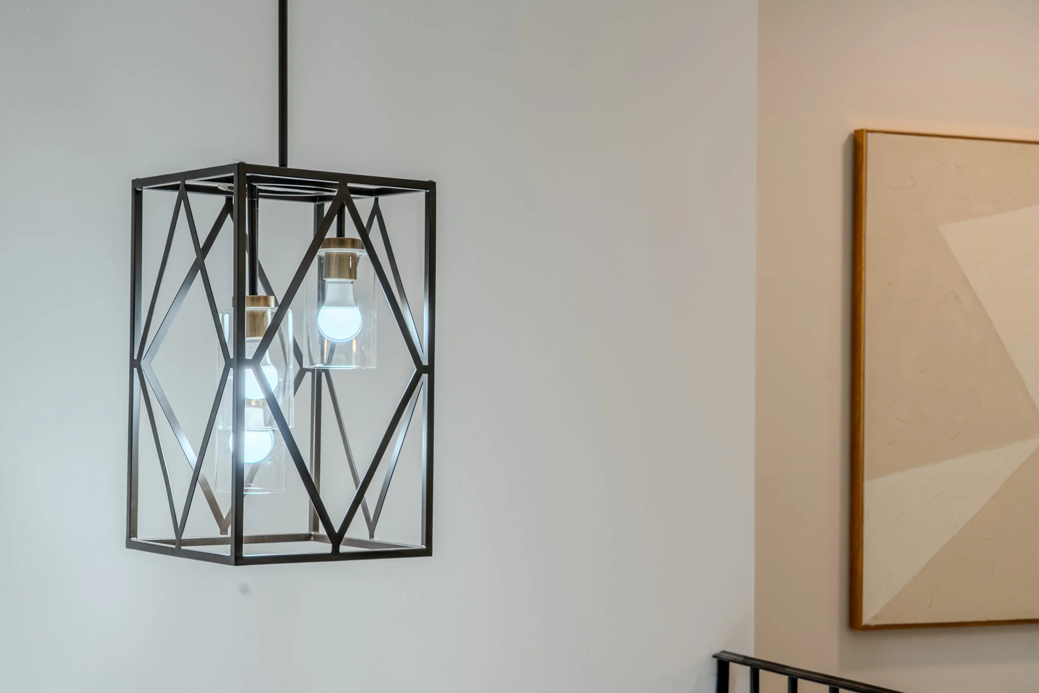 A modern wall-mounted black metal light fixture with multiple exposed bulb lights, next to a beige wall with a framed abstract art piece.