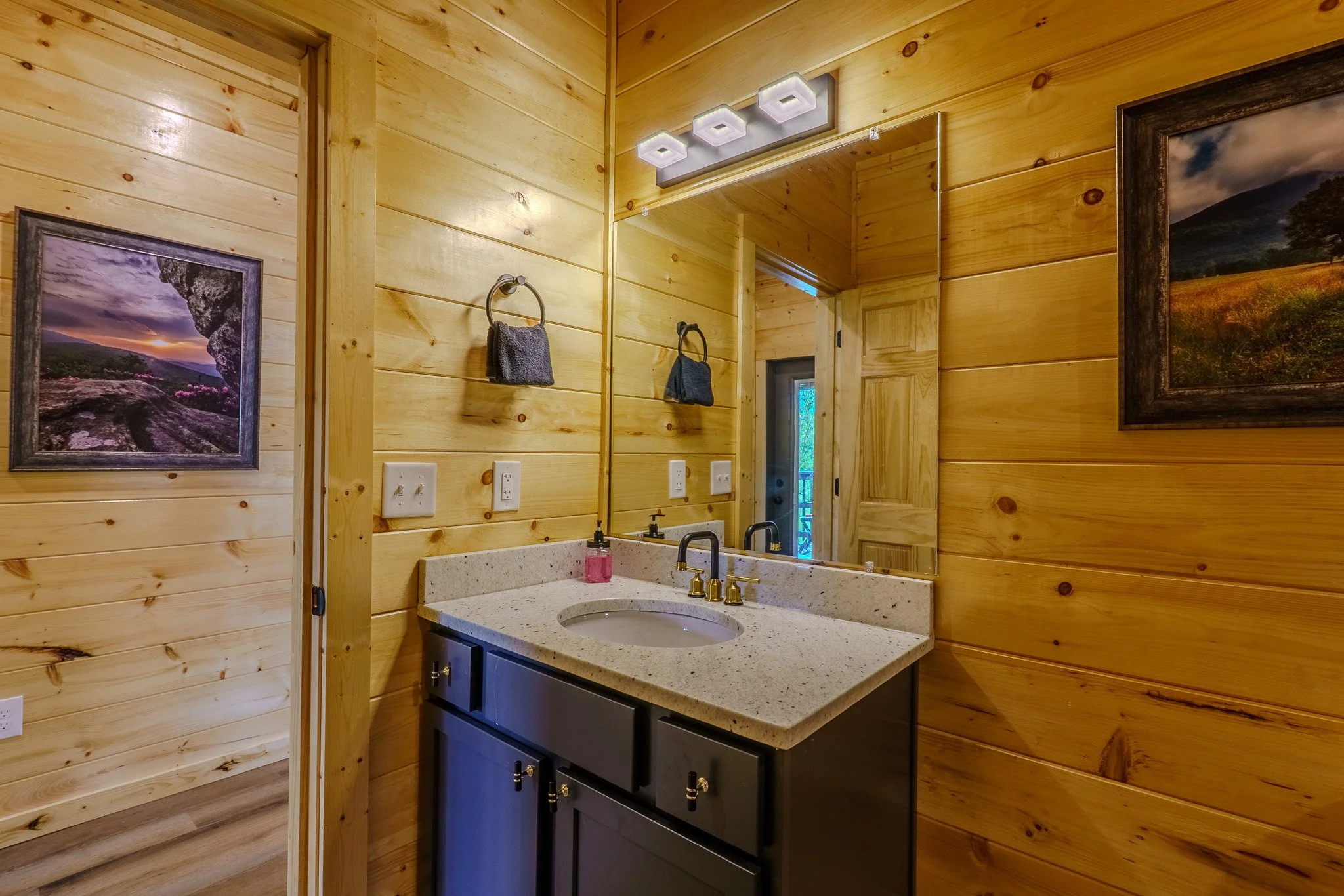 Bathroom with wooden walls, black vanity with a granite countertop, large mirror, wall-mounted light fixture, towels hanging, framed landscape photos, door leading outside.