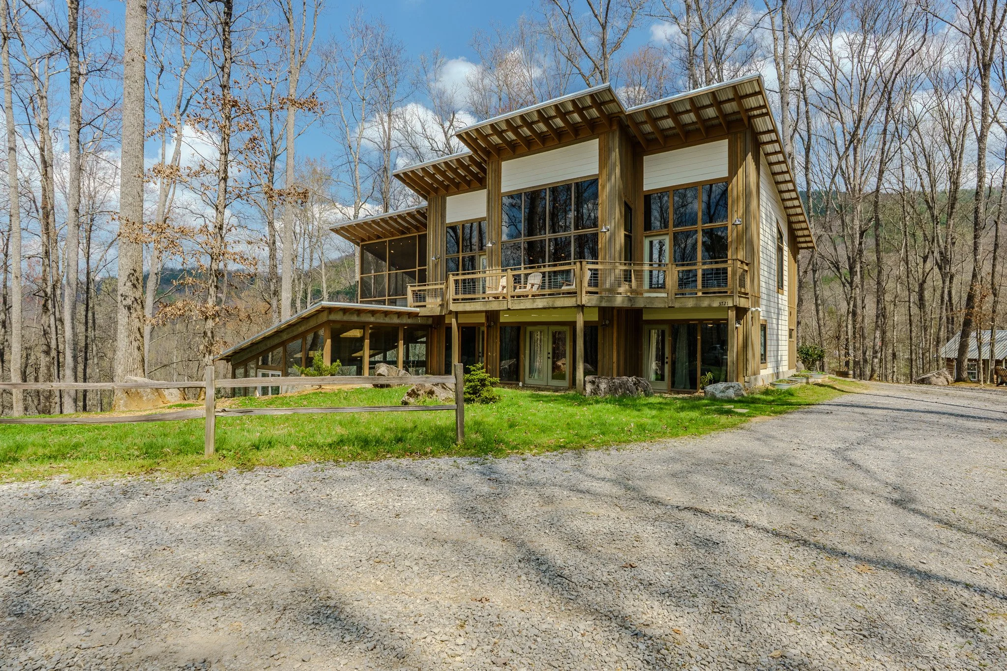 A modern multi-story house with large windows and a wooden exterior, situated in a wooded area. It has multiple decks and a gravel driveway in front.