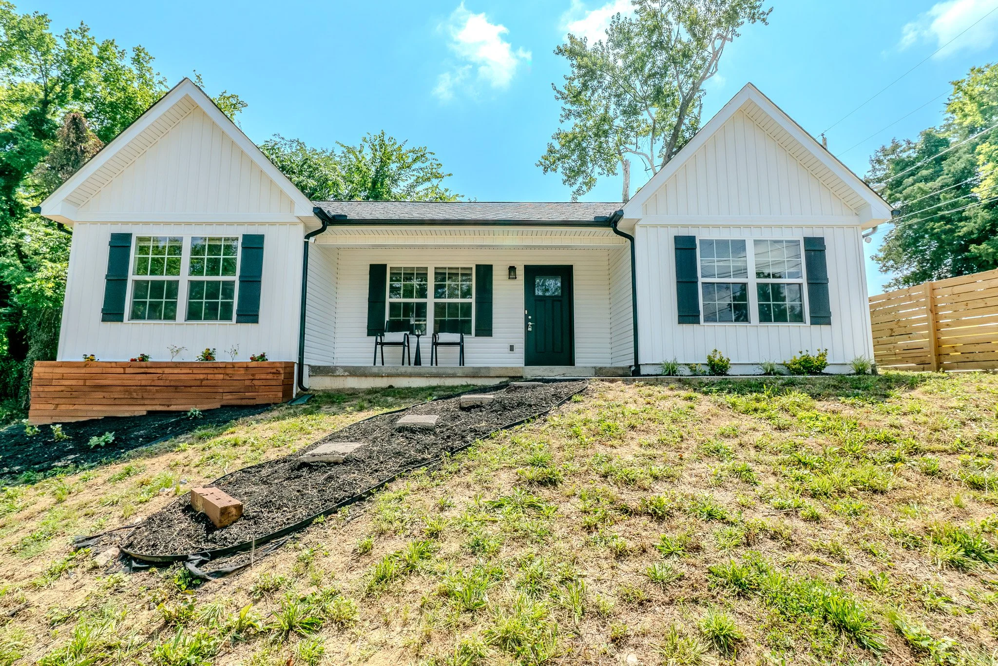 White house with black shutters, front porch with two chairs, black door, on a grassy hillside with a new wooden planter box and wooden fence, under a blue sky with trees in the background.
