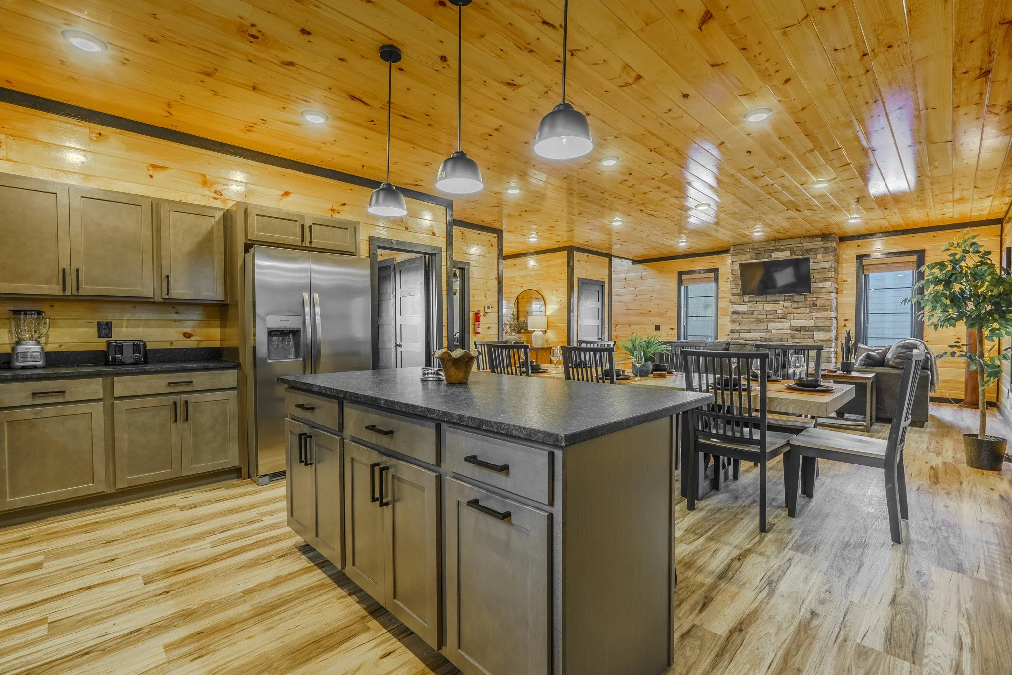Open-concept kitchen and dining area with wood-paneled walls and ceiling, featuring a central island with a black countertop, a stainless steel refrigerator, a wall-mounted TV, and a stone fireplace in the background, with windows allowing natural li