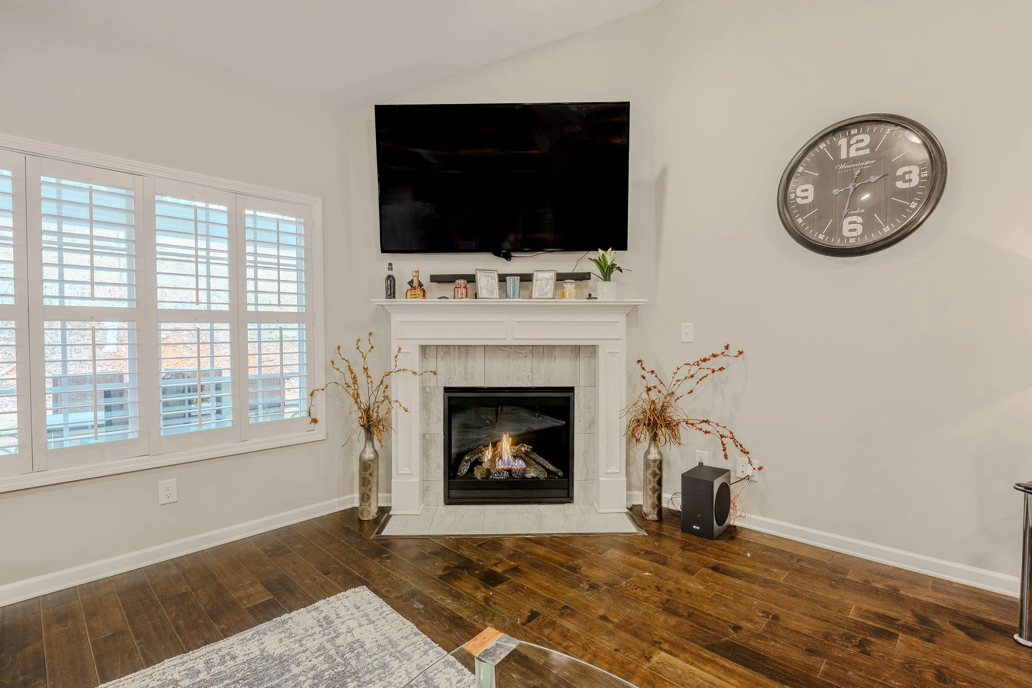 Living room with a fireplace, wall-mounted flat-screen TV, vases with dried branches, and a large wall clock.