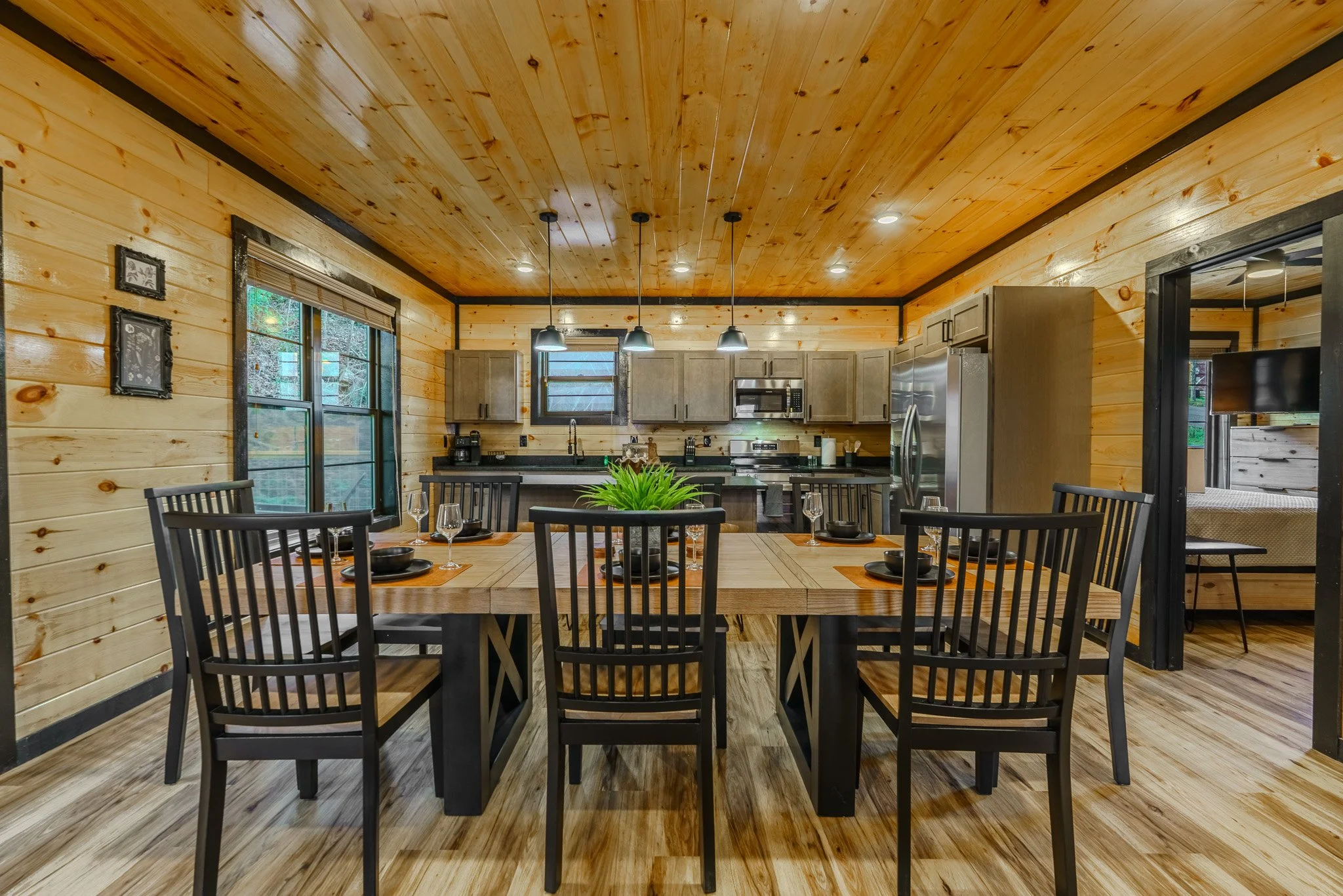 Interior of a wooden cabin-style dining area with a large wooden table, black chairs, and a kitchen in the background with gray cabinets and stainless steel appliances.