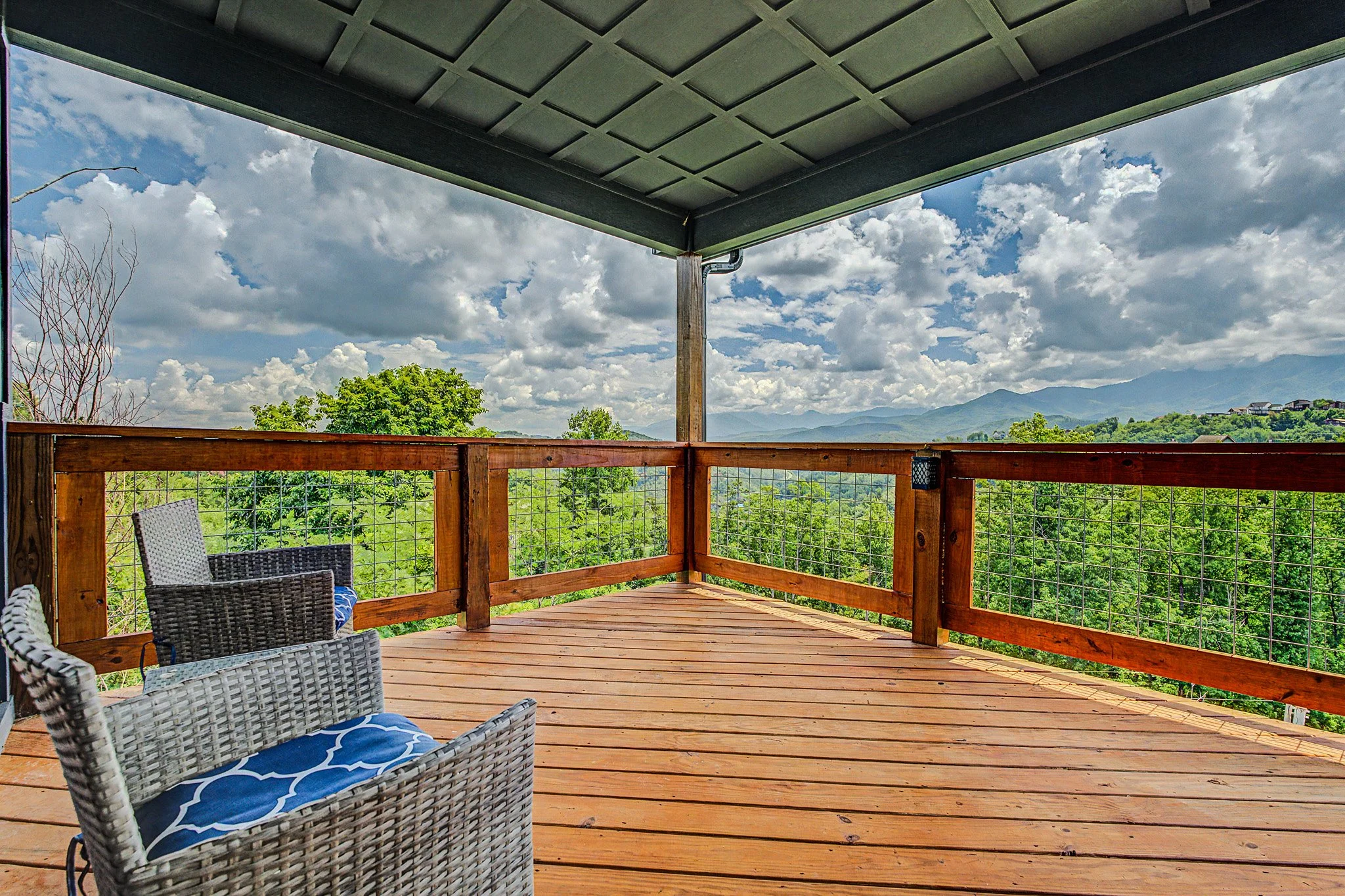 View of a wooden deck with outdoor furniture, overlooking trees and distant mountains under a partly cloudy sky.