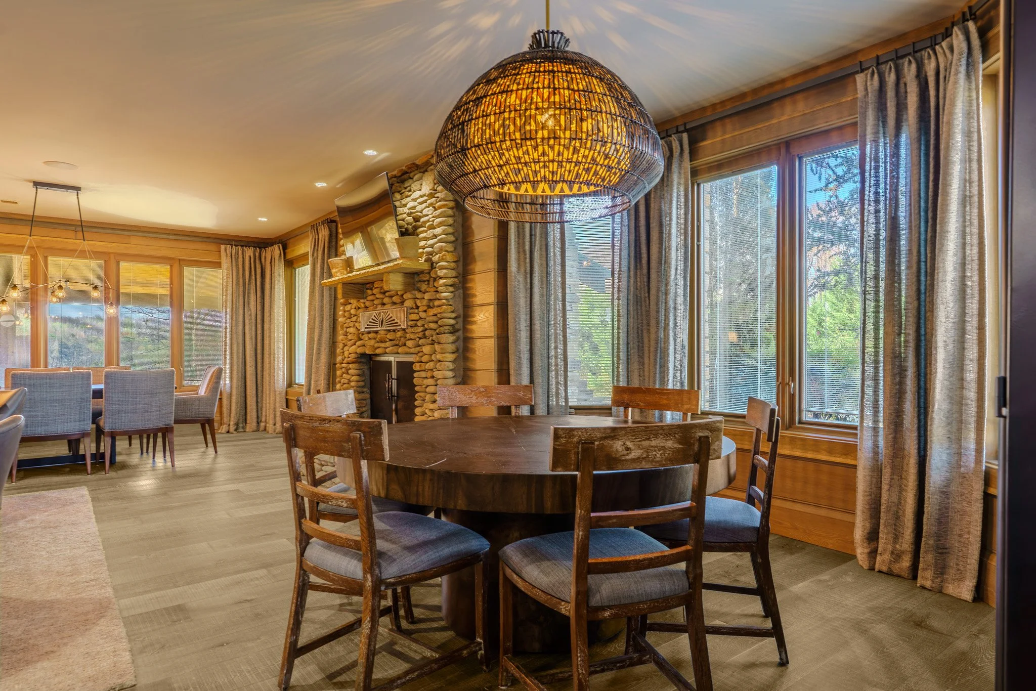 A cozy dining area with a round wooden table and six chairs, large windows with curtains, a stone fireplace, and a rattan chandelier hanging from the ceiling.