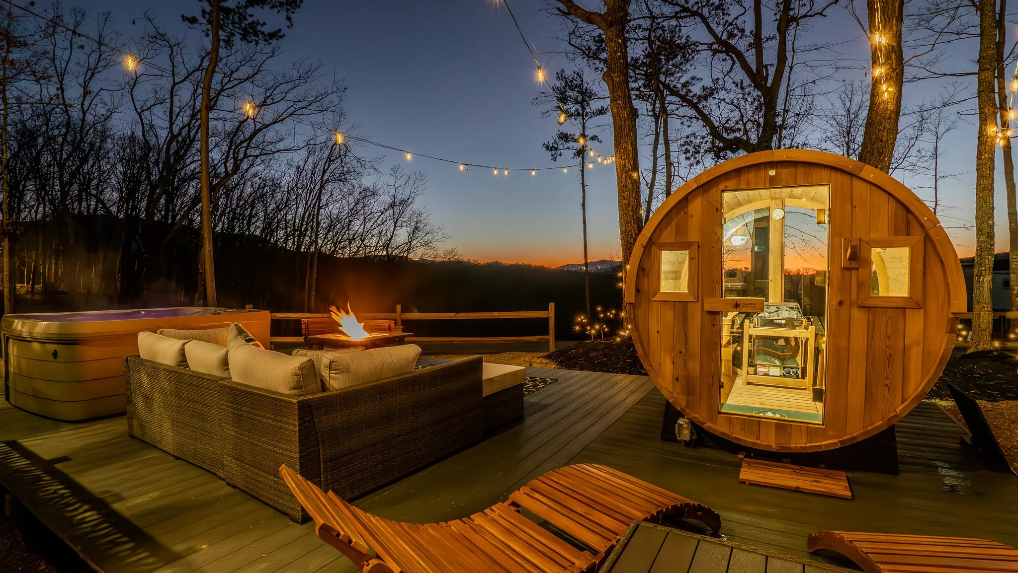 Outdoor scene at dusk featuring a wooden barrel-shaped sauna, a hot tub, a cushioned outdoor sofa, a fire pit, string lights, and a scenic view of trees and mountains.