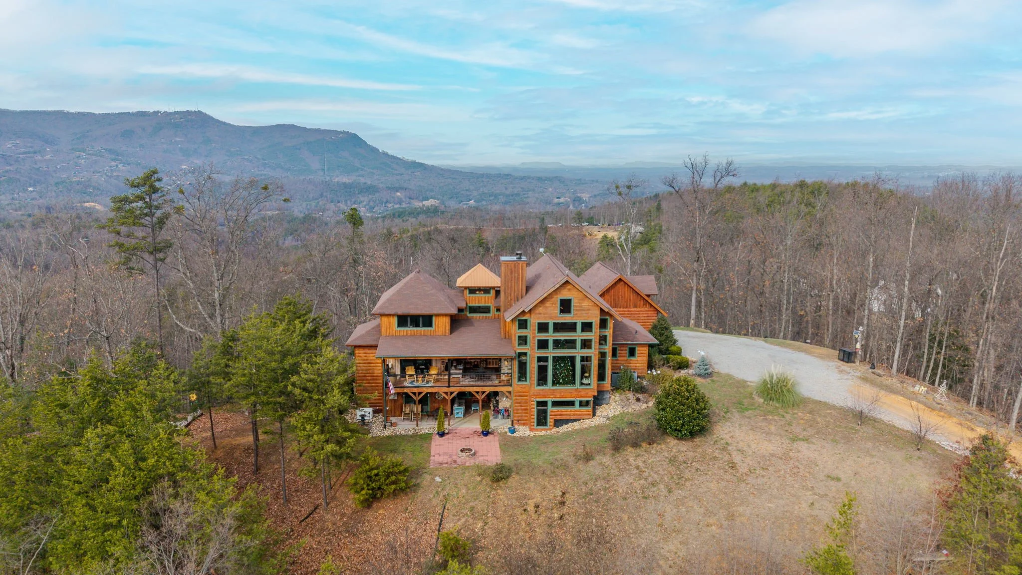 A large wooden house on a hill surrounded by trees and overlooking a mountainous landscape, with a gravel driveway and garden area.