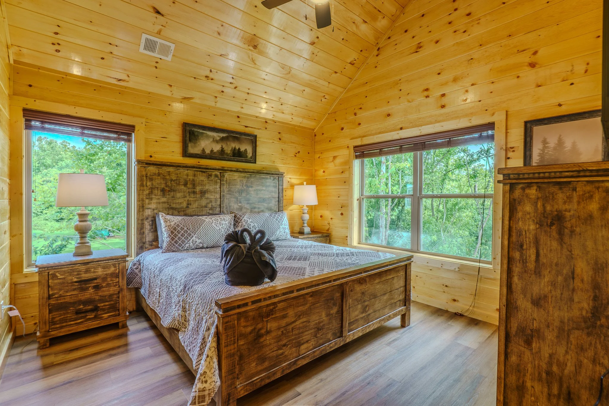 A cozy bedroom with wooden walls, ceiling, and floor, featuring a large bed with patterned bedding, a wooden headboard, two bedside tables with lamps, and large windows showing green trees outside.