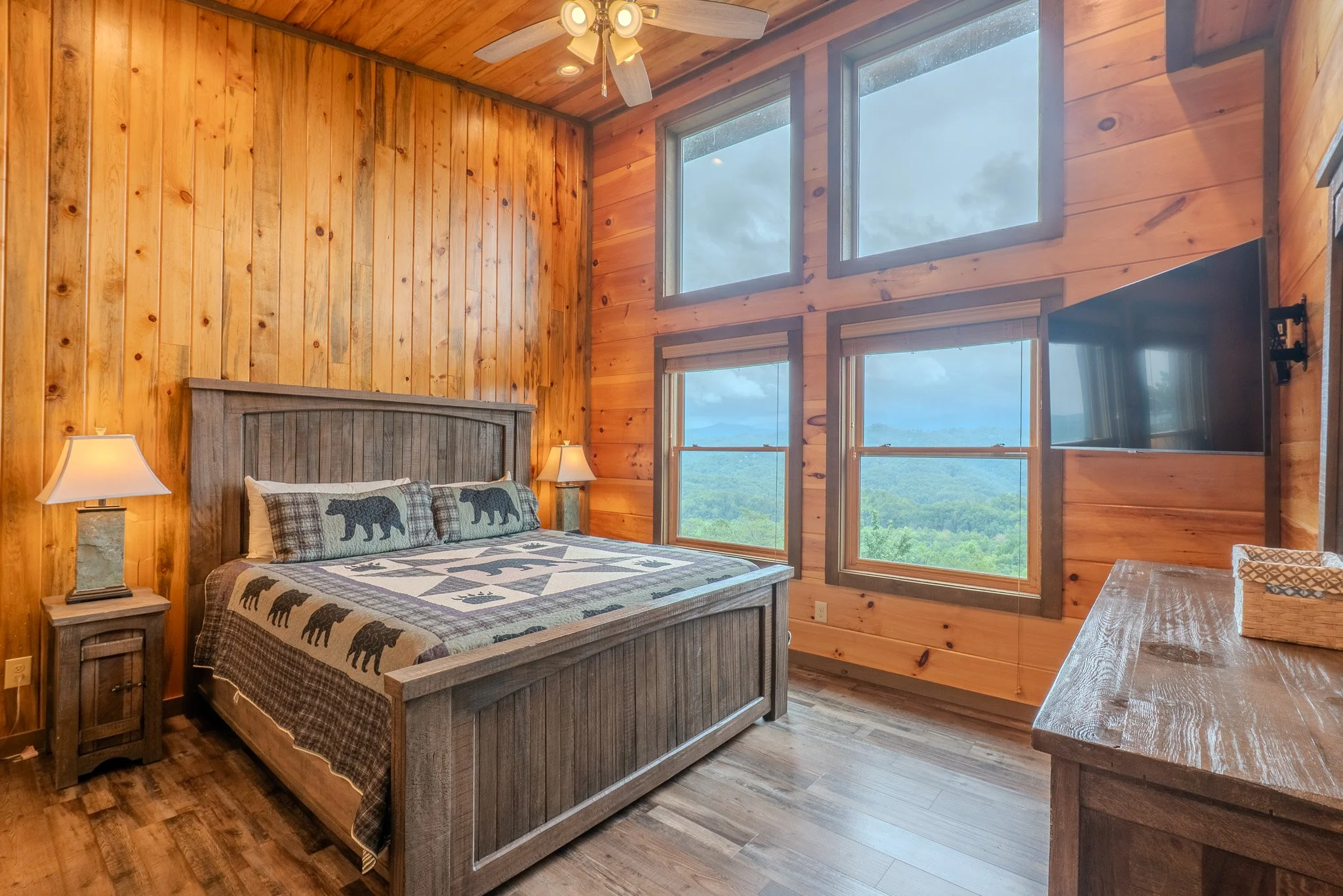 Wood-paneled bedroom with large windows showing a scenic view of mountains and trees, rustic wooden furniture, a bed with bear-themed quilt and pillows, and a flat-screen TV mounted on the wall.