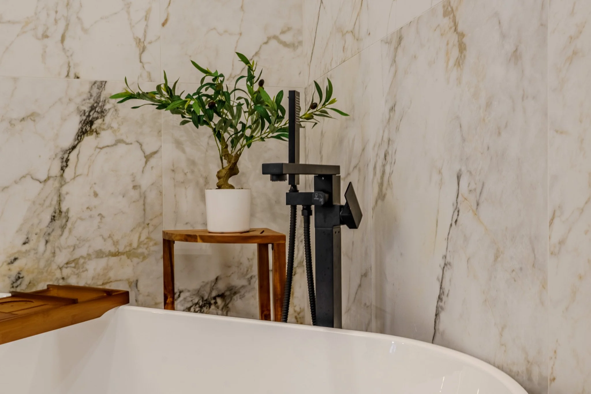 A bathtub with a wooden side panel, a small wooden table holding a potted green plant, and a black bathtub faucet mounted on a marbled wall.