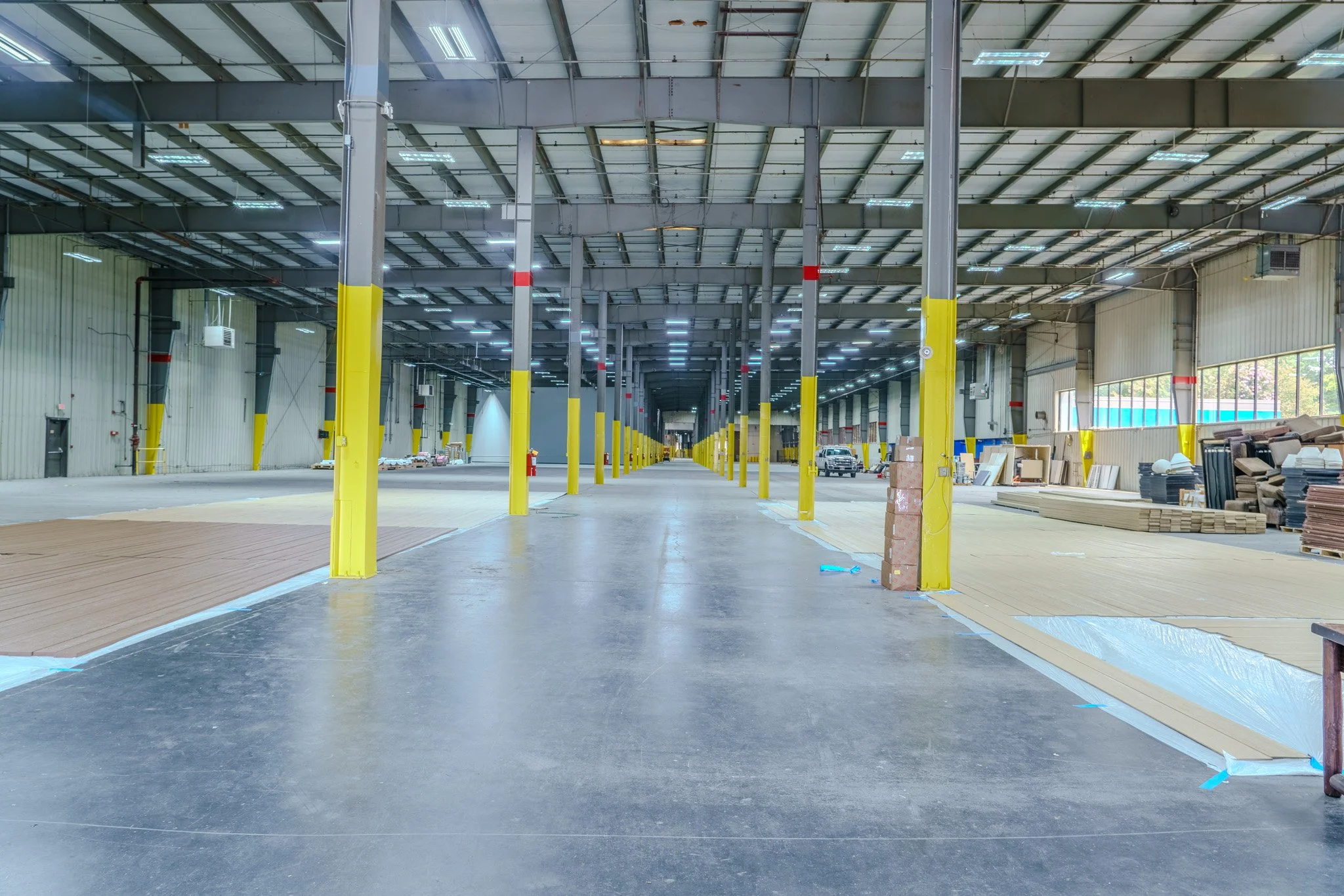 Interior of a large warehouse or industrial space with yellow support beams, stacked materials on the sides, and a wide open aisle.