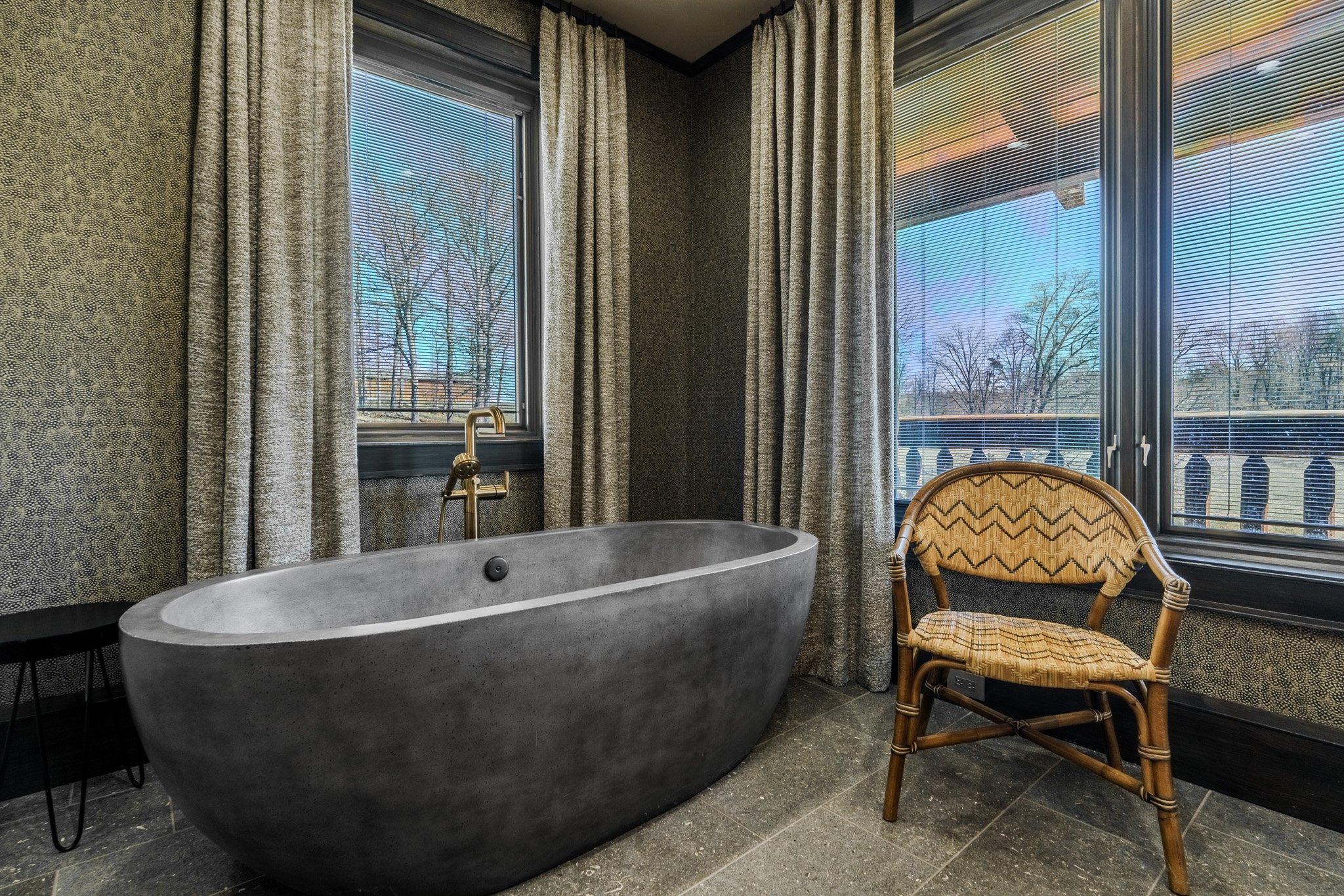 A modern bathroom with a freestanding gray bathtub, a large window with curtains showing a landscape, and a wicker chair with patterned cushions.