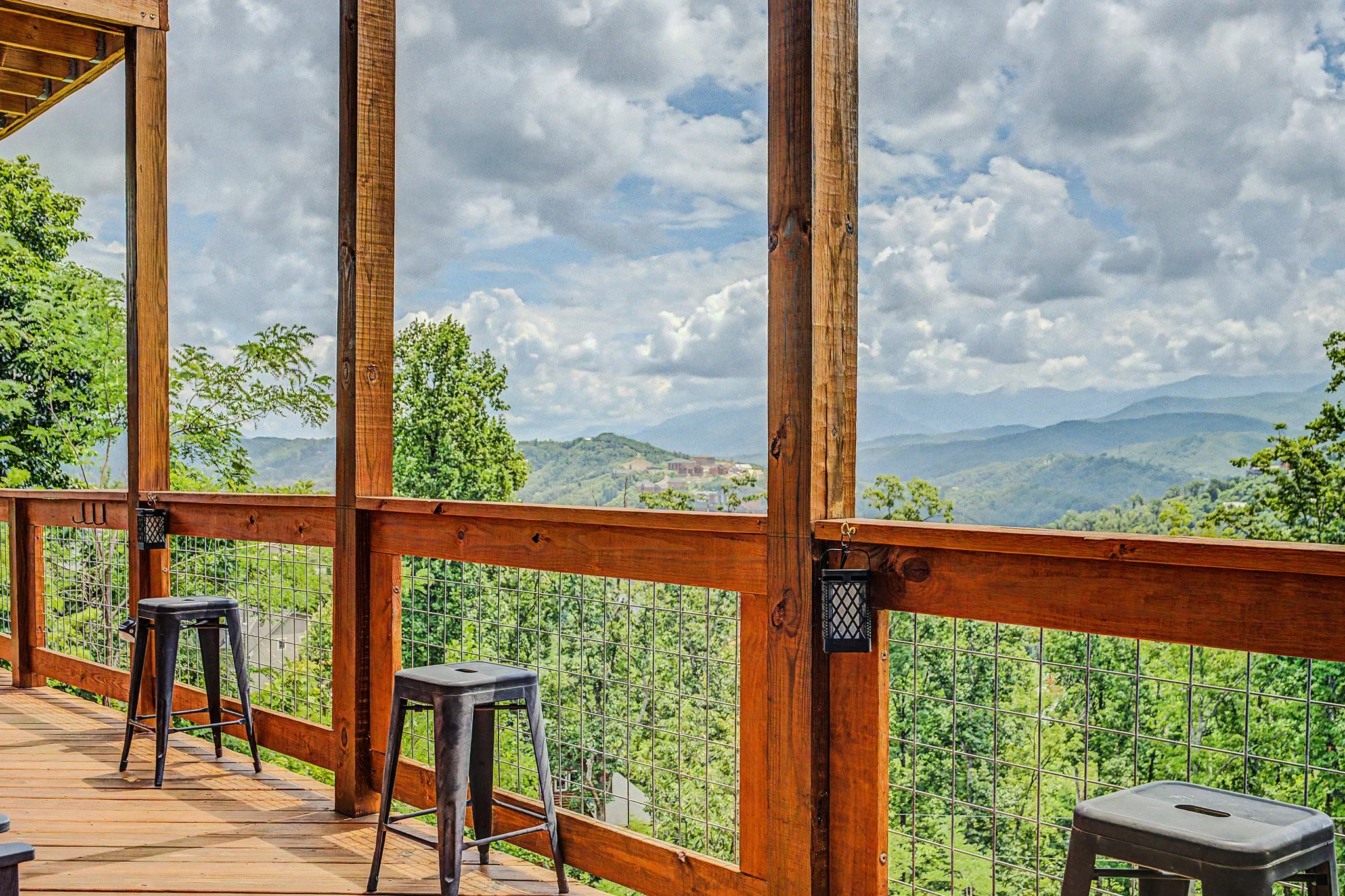 A partially constructed wooden balcony or deck with scenic mountain view in the background. There are two black stools, a plastic storage container, and two lanterns hanging on the wooden railing.
