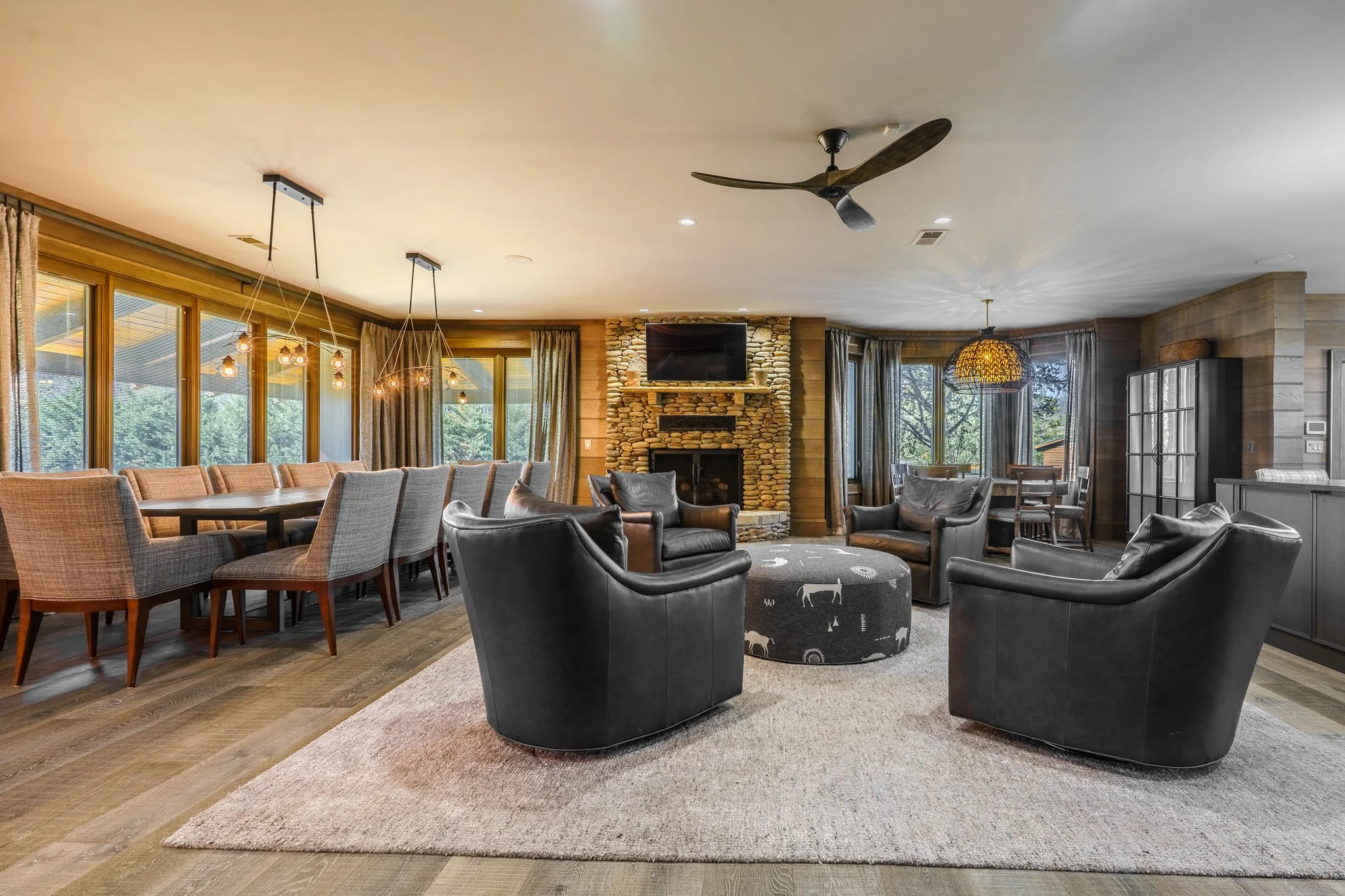 Open-concept living and dining room with large windows, a stone fireplace, four black leather armchairs arranged around a round ottoman with animal print, and a long dining table with upholstered chairs, warm wooden flooring, and modern lighting.