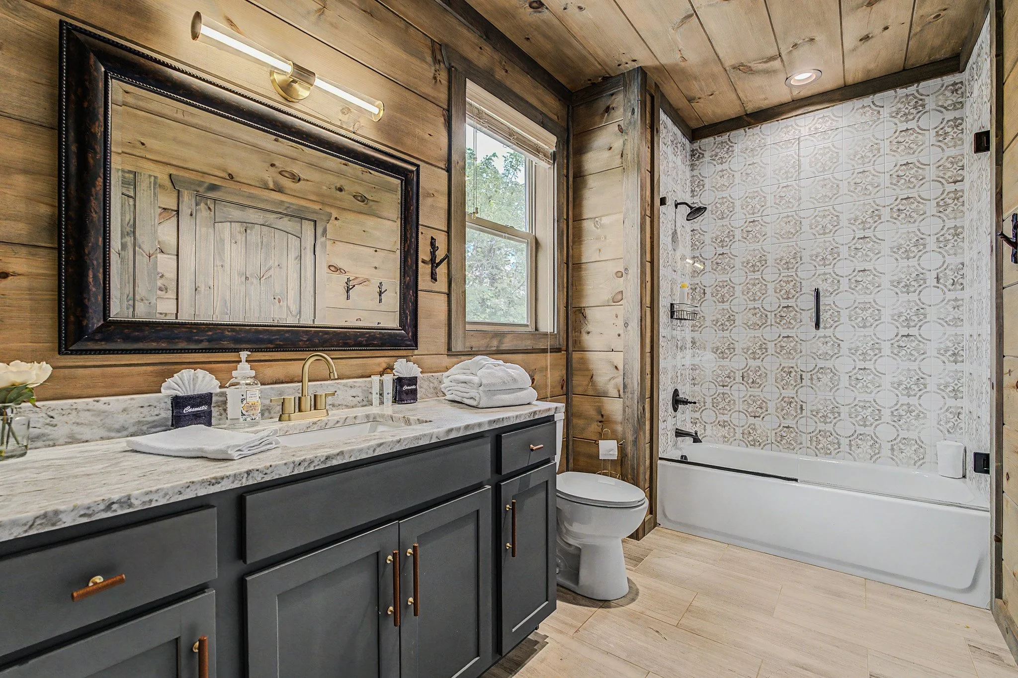 Bathroom featuring a vanity with a marble countertop, black cabinets with gold handles, a large mirror, and a wall-mounted light fixture. There is a window, wooden paneled walls, and a shower area with patterned tiles, a showerhead, corner shelf, and