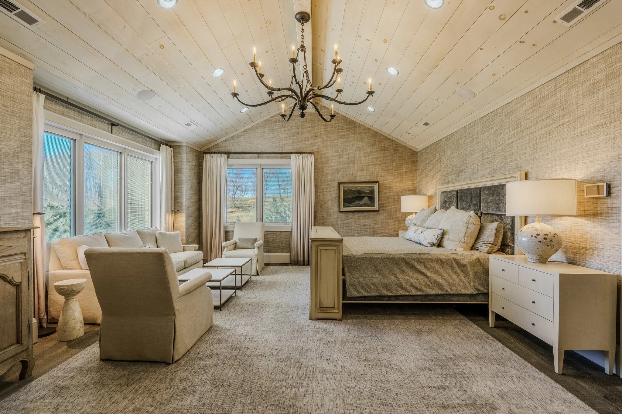 A spacious bedroom with a high wooden ceiling, a chandelier, a large window with curtains, a bed with multiple pillows, and a sitting area with a sofa, armchair, and coffee tables.