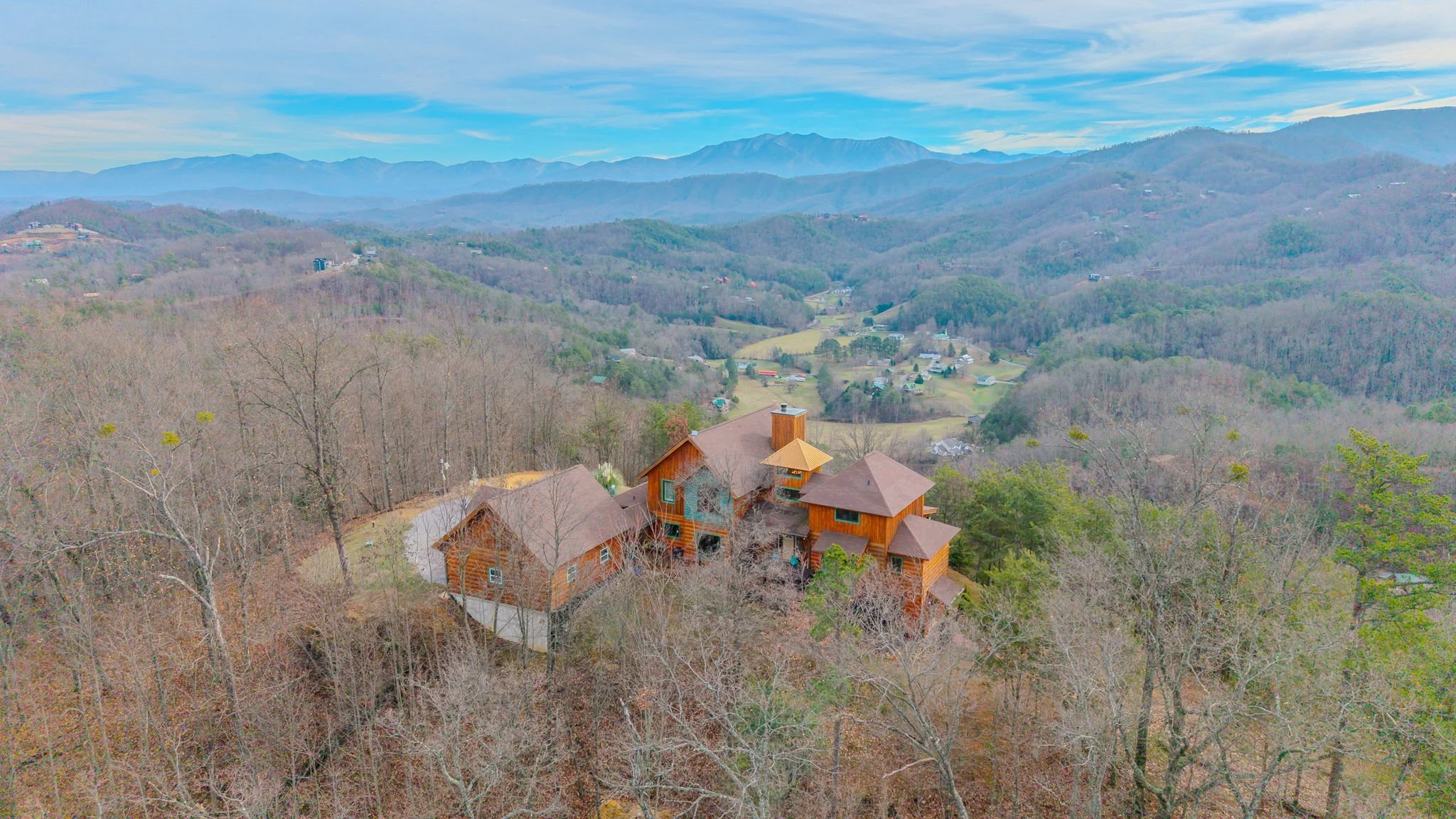 Aerial view of a large wooden house in a hilly, forested area with mountains in the background, under a partly cloudy sky.
