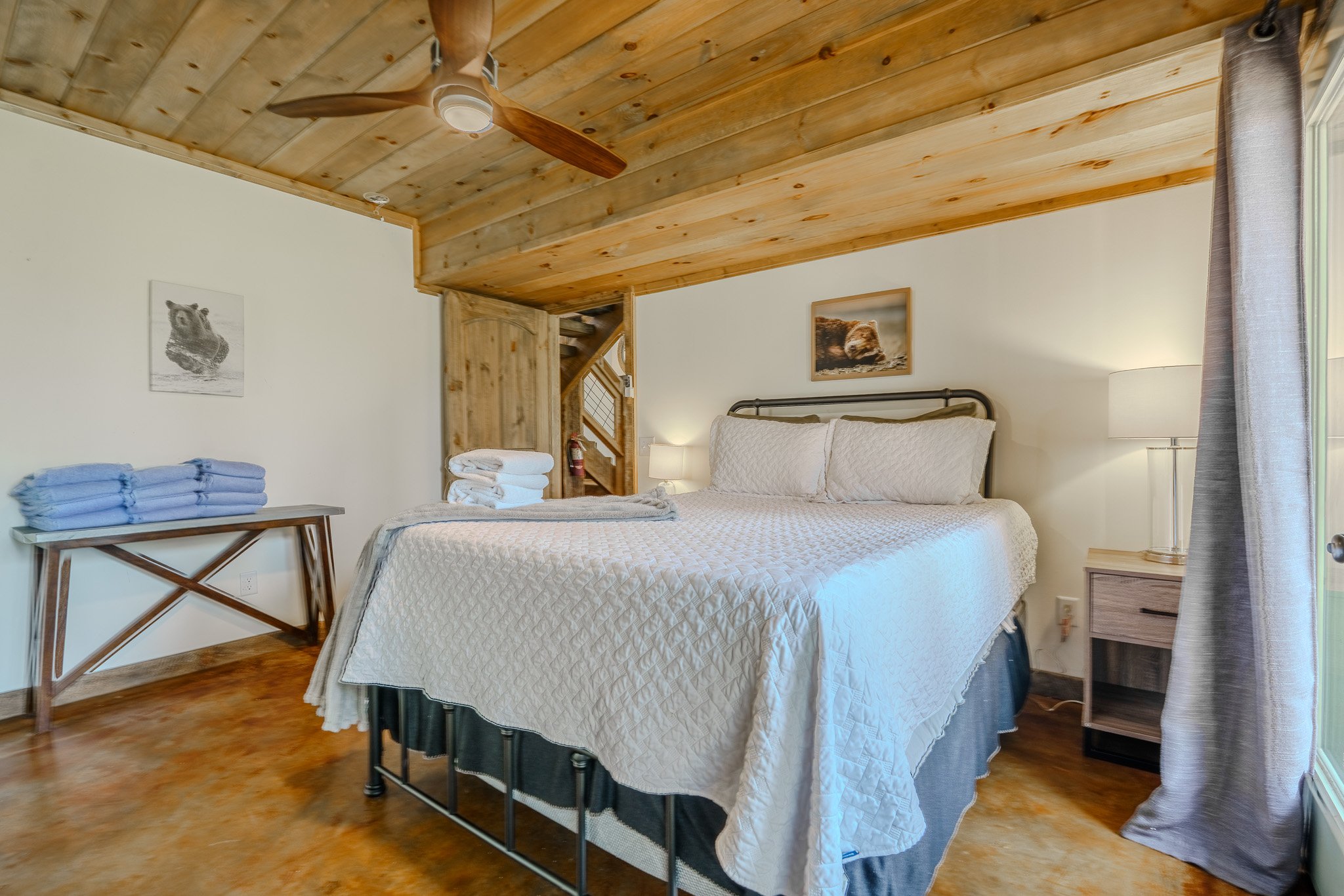 Bedroom with a wooden ceiling and floor, a bed with white bedding, a nightstand with a lamp, a window with gray curtains, and pictures of bears on the wall.