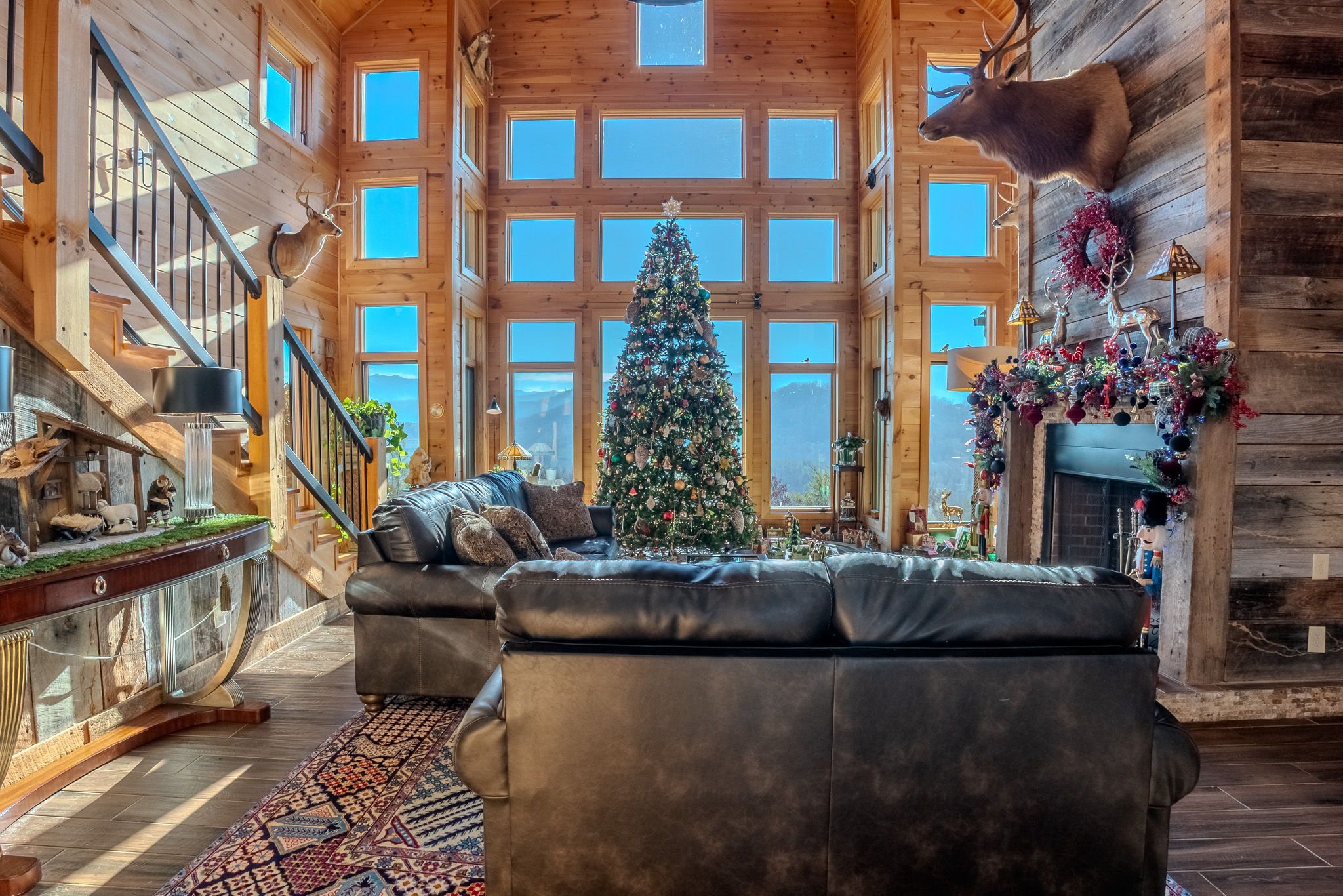 A cozy living room with wooden walls, decorated for Christmas with a large Christmas tree in the center. The room features a high ceiling with many windows showing a mountain landscape. There are mounted deer head decorations on the walls, a fireplac
