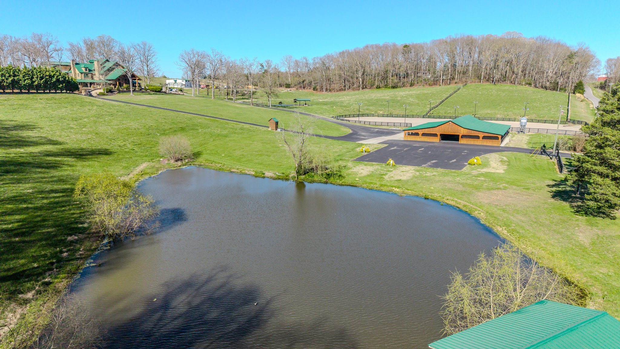 A park with a small pond, green lawns, trees, and a building with a green roof and a parking lot.