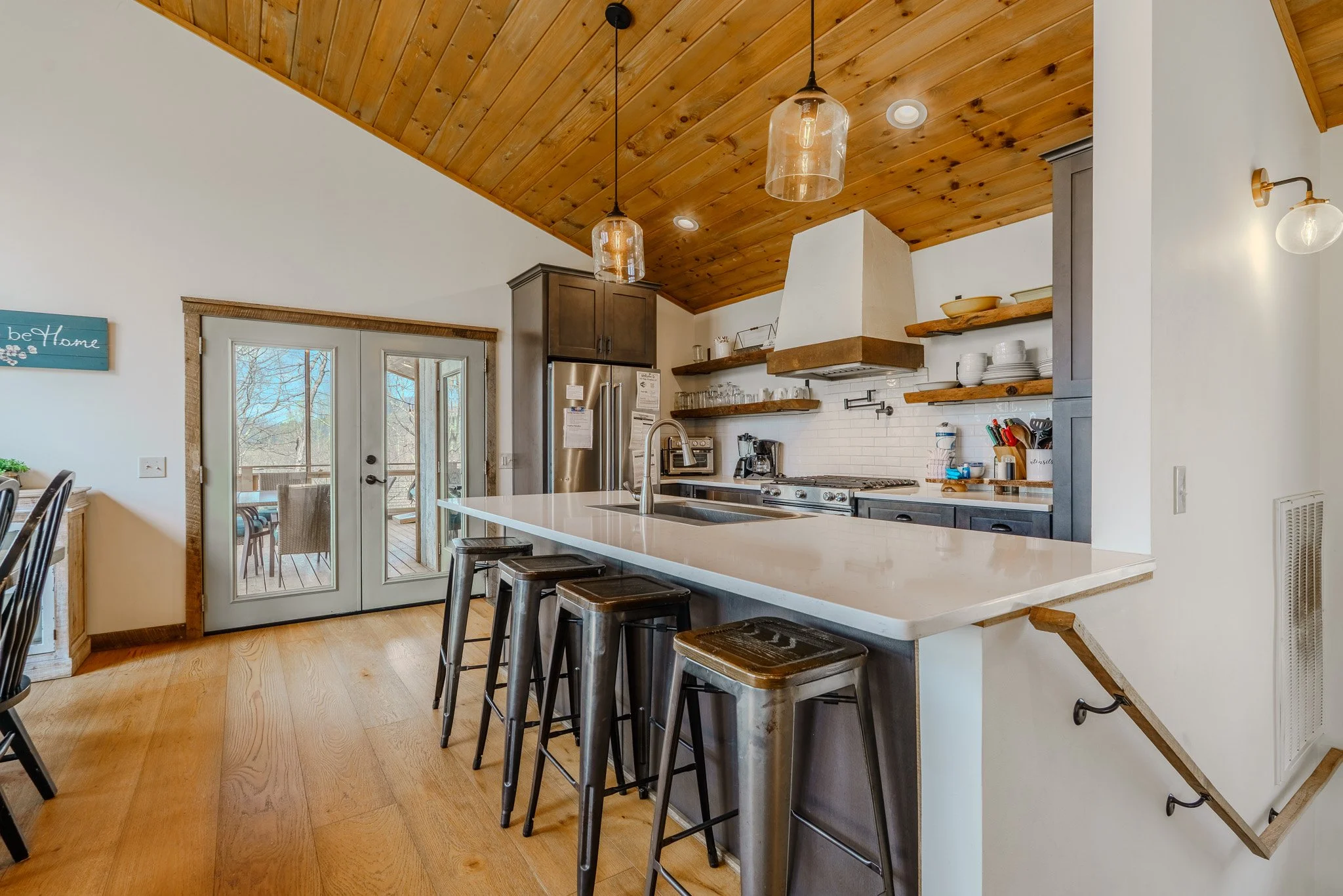 Modern kitchen with a white island countertop surrounded by bar stools, stainless steel appliances, open wooden shelves, pendant lights, a sliding glass door leading to an outdoor deck, and a vaulted wooden ceiling.
