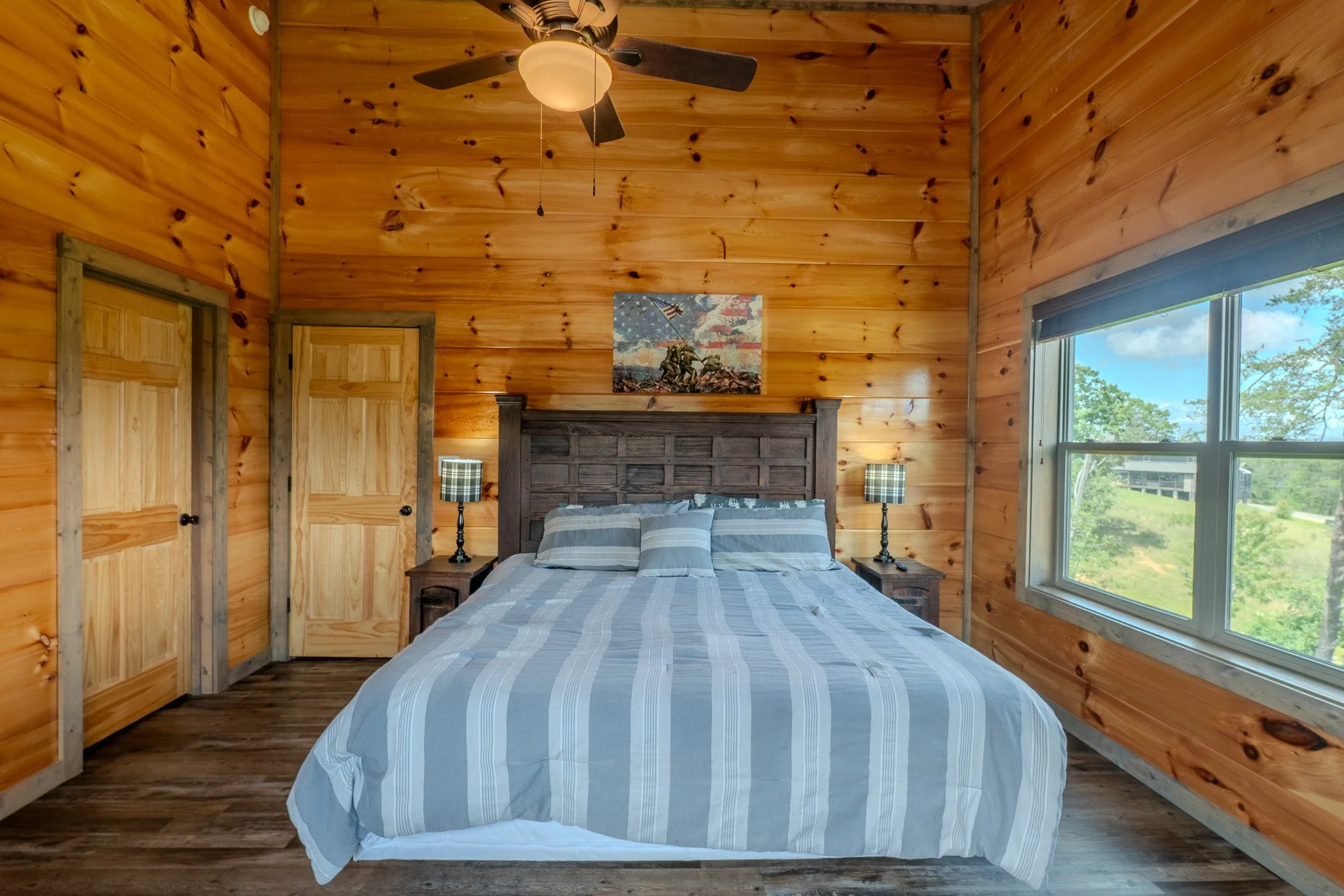 A cozy bedroom with wood-paneled walls and ceiling, featuring a large bed with striped bedding, two bedside tables with plaid lamps, a ceiling fan, a window showing green trees outside, and a mountain landscape painting above the bed.