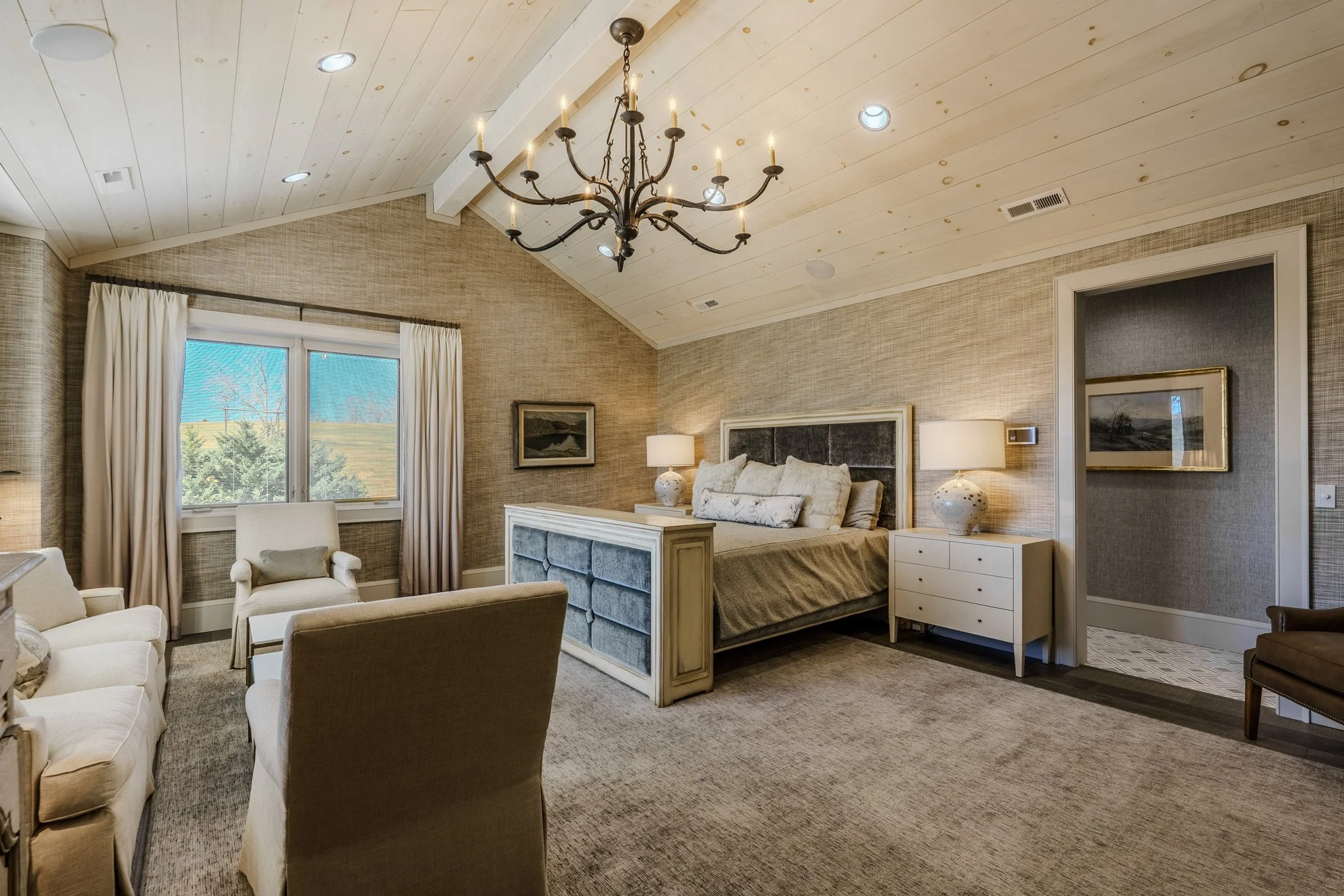A bedroom with a vaulted ceiling, a chandelier, a window with beige curtains, a bed with pillows, nightstands with lamps, a sitting area with chairs and a sofa, and framed artwork on the walls.
