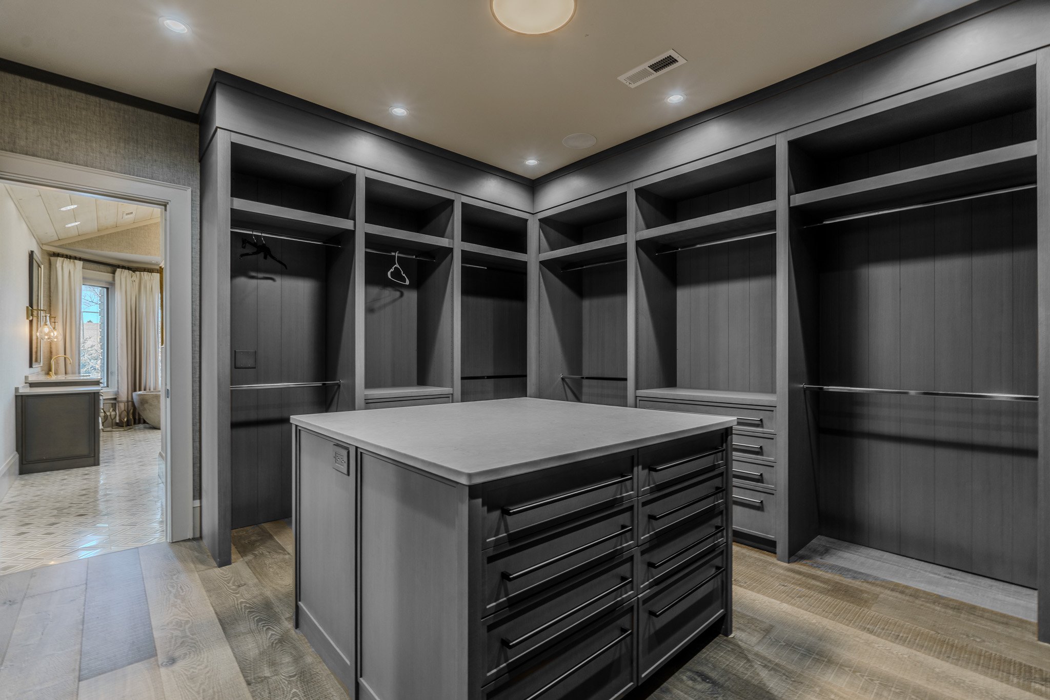 Empty walk-in closet with dark gray cabinetry, open shelves, and drawers, with a small island in the center, and an adjoining room visible through a door on the left.