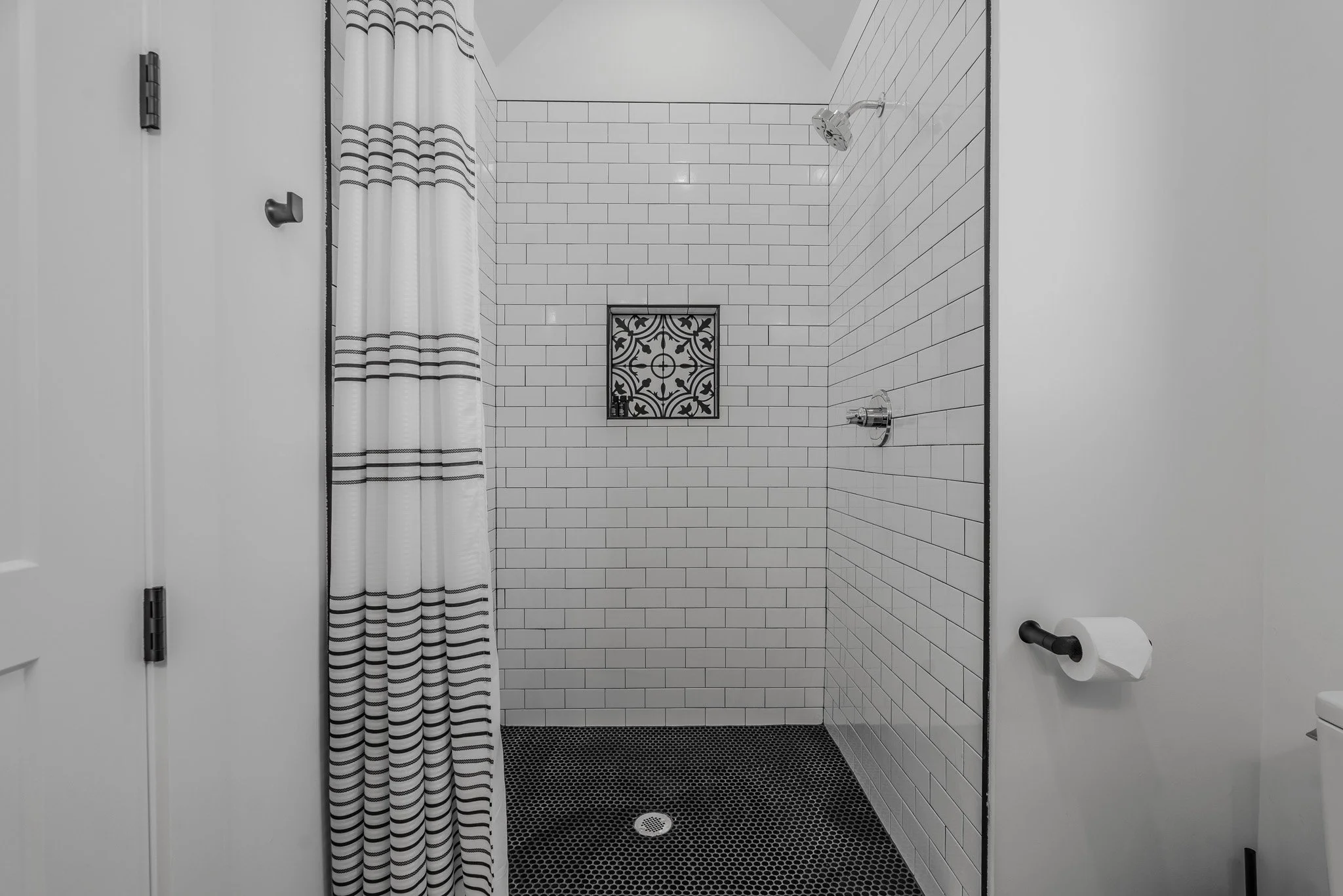Empty walk-in shower with white subway tiles, black hexagonal floor tiles, a decorative black and white tile accent in the wall, a silver showerhead, and a striped shower curtain.