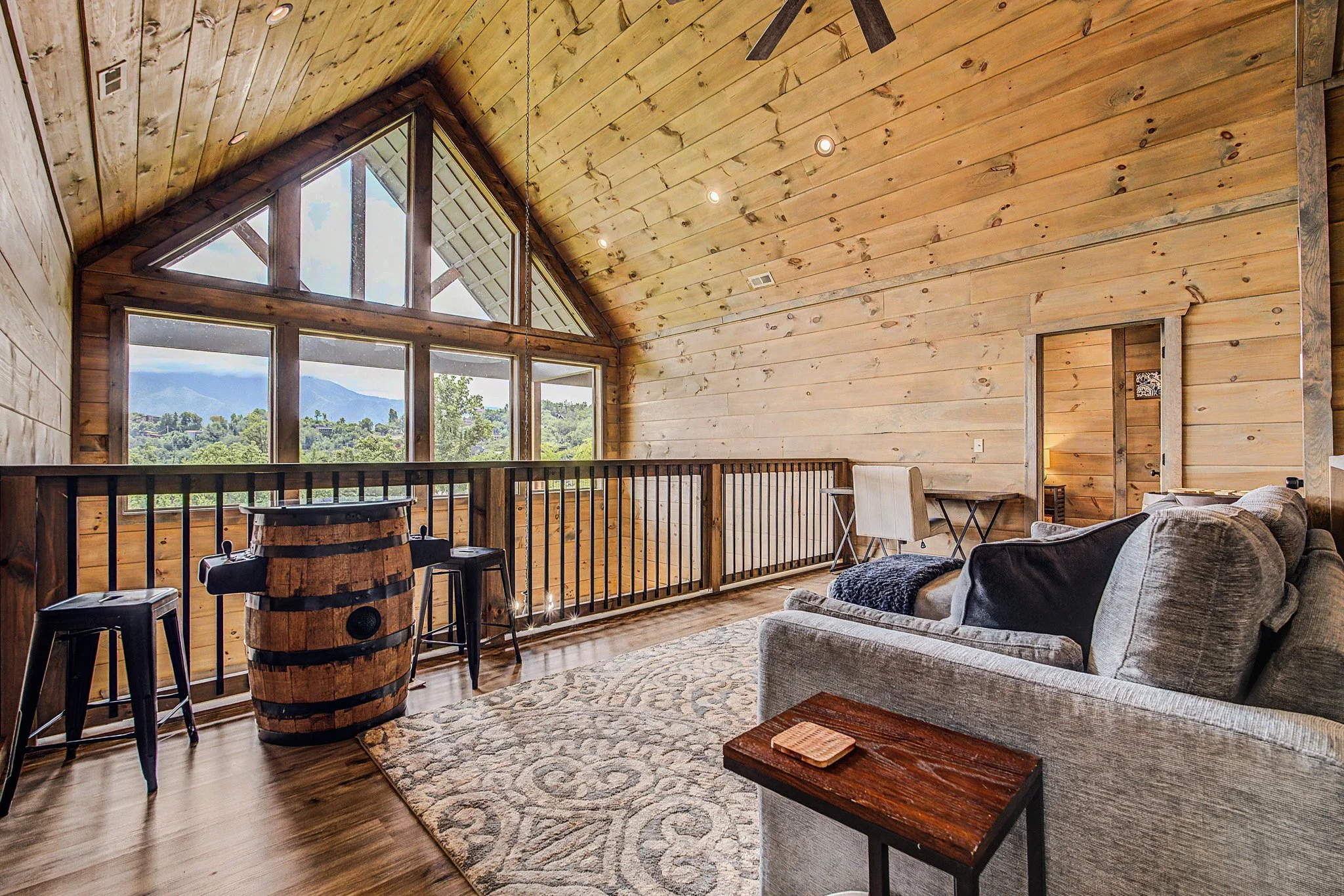 Rooftop living area with wooden walls and ceiling, large triangular windows showing mountains and trees outside, a sofa, a small table, and a barrel-shaped wine rack with black stools nearby.