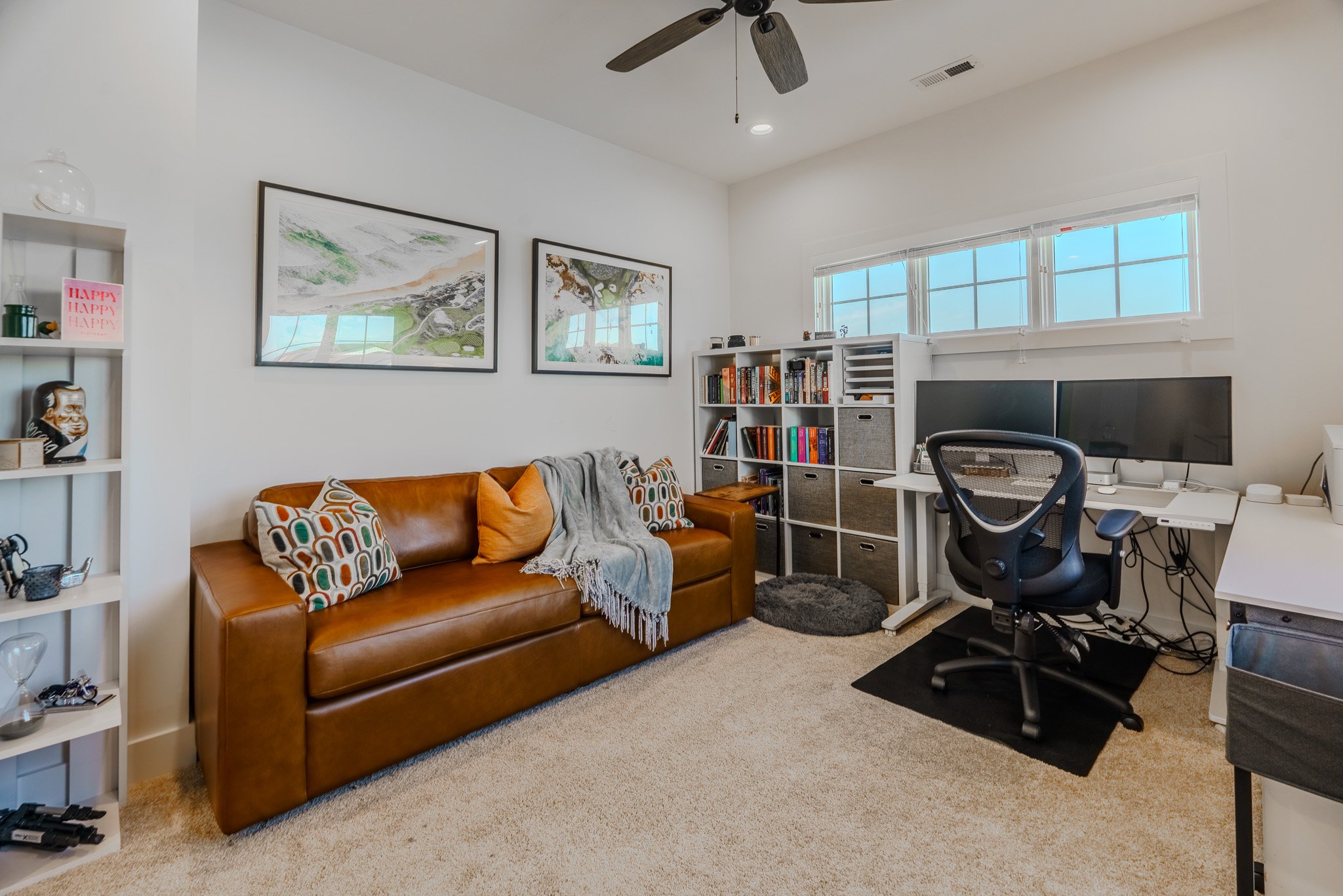 A cozy home office with a brown leather sofa, decorative pillows, a gray blanket, and a white bookshelf filled with books and decor. There is a dual monitor desk with an office chair, and two horizontal windows above it, letting in natural light.