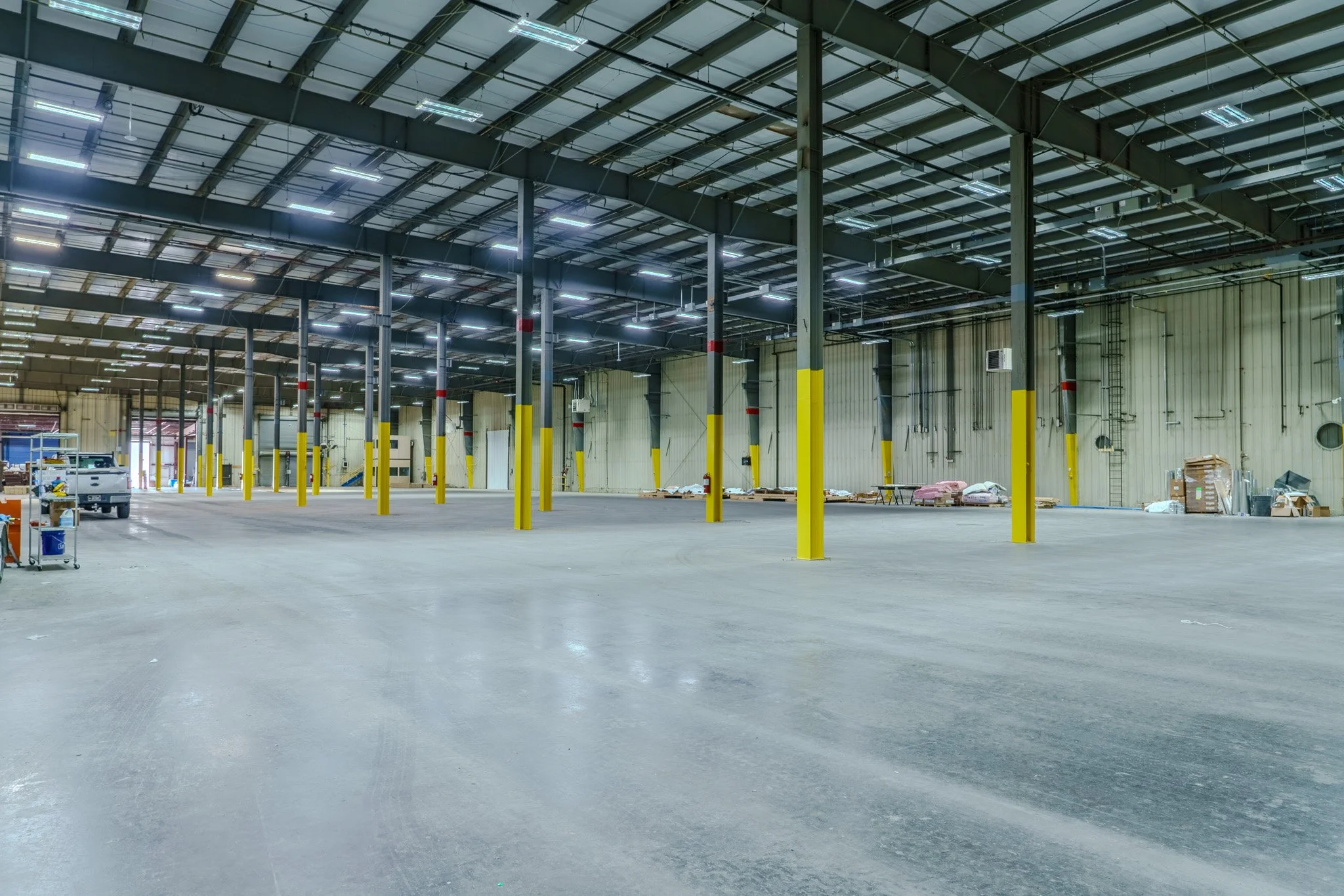 Empty warehouse with yellow safety posts, a truck on the left, and construction materials along the back wall.