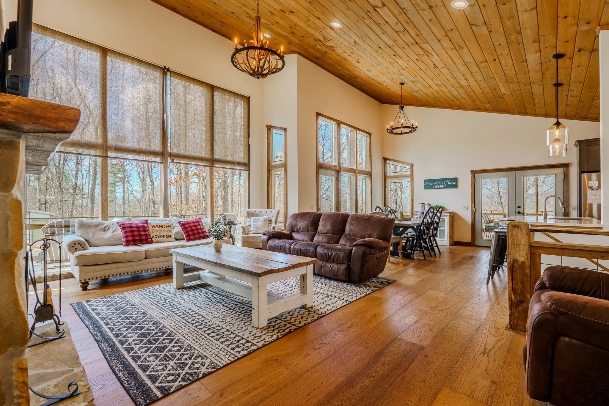 Spacious living room with large windows, wooden ceiling, and hardwood floors. It features a white sofa with red plaid pillows, a brown leather sofa, a rustic coffee table, a patterned rug, and a dining area with black chairs. Natural light fills the 