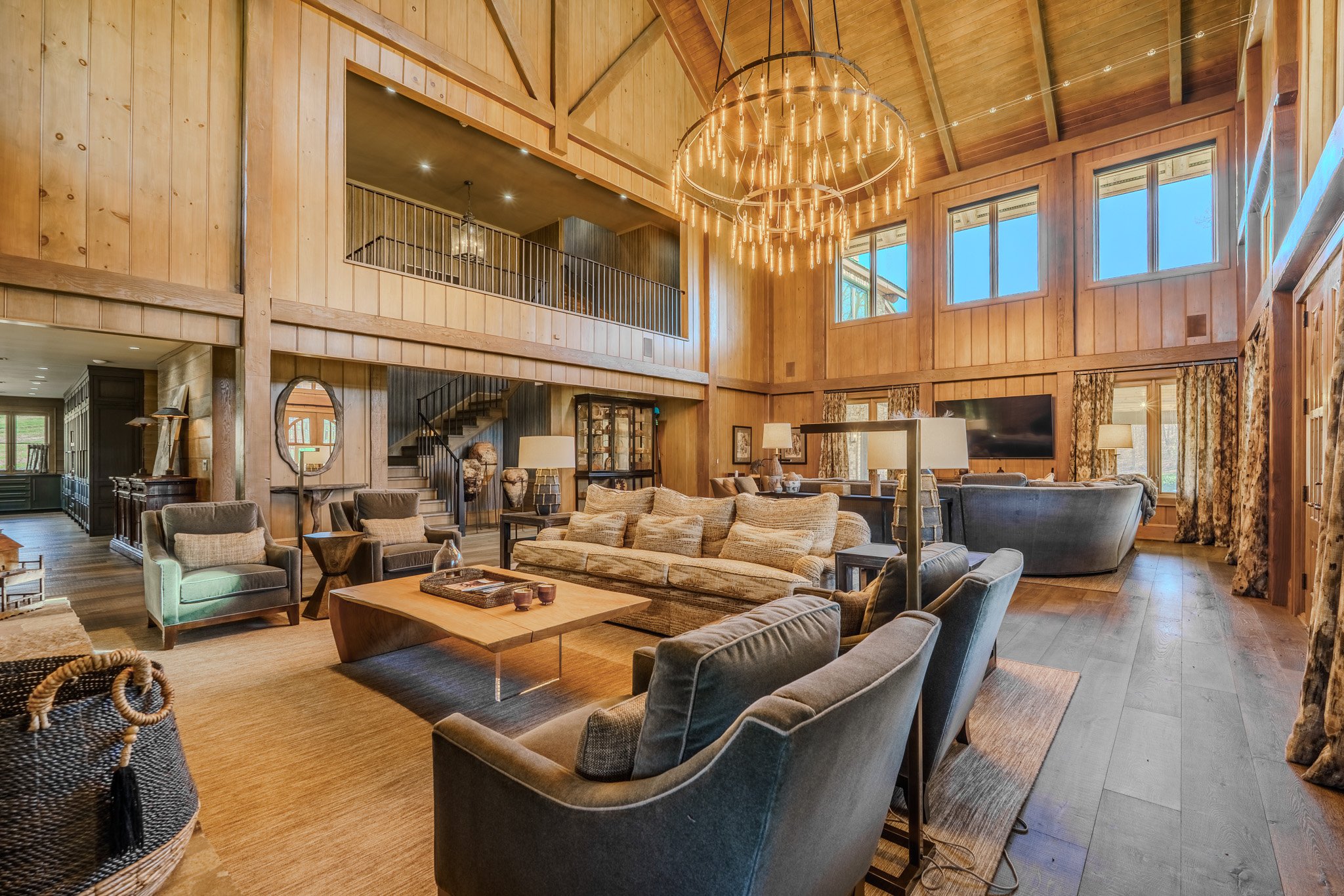 A spacious living room with tall wooden walls and ceiling, featuring multiple windows, a large chandelier, sofas, armchairs, a television, and decorative lamps.